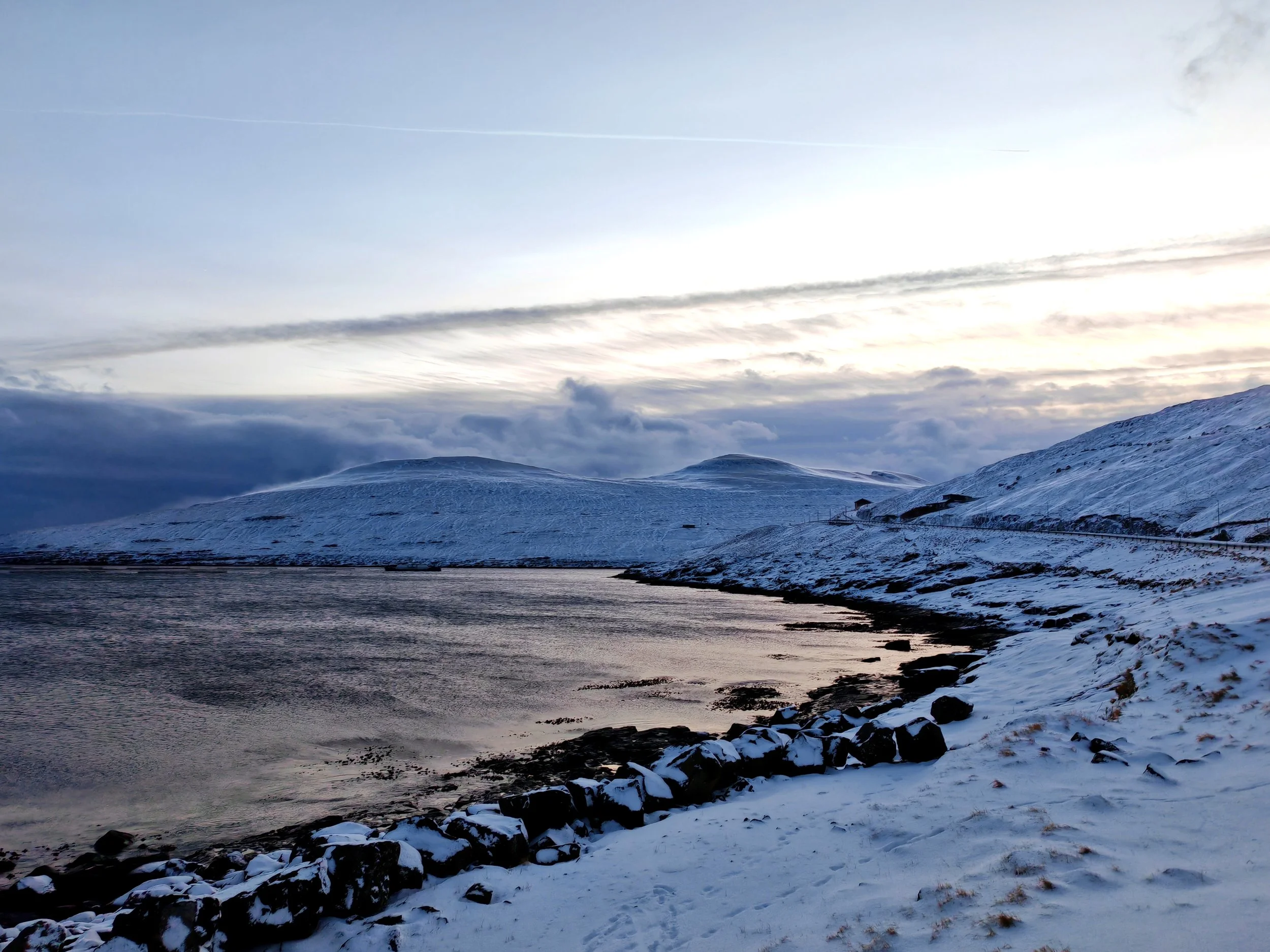 Snow-covered mountains near a body of water with a cloudy sky and a few structures at the base of the mountains.