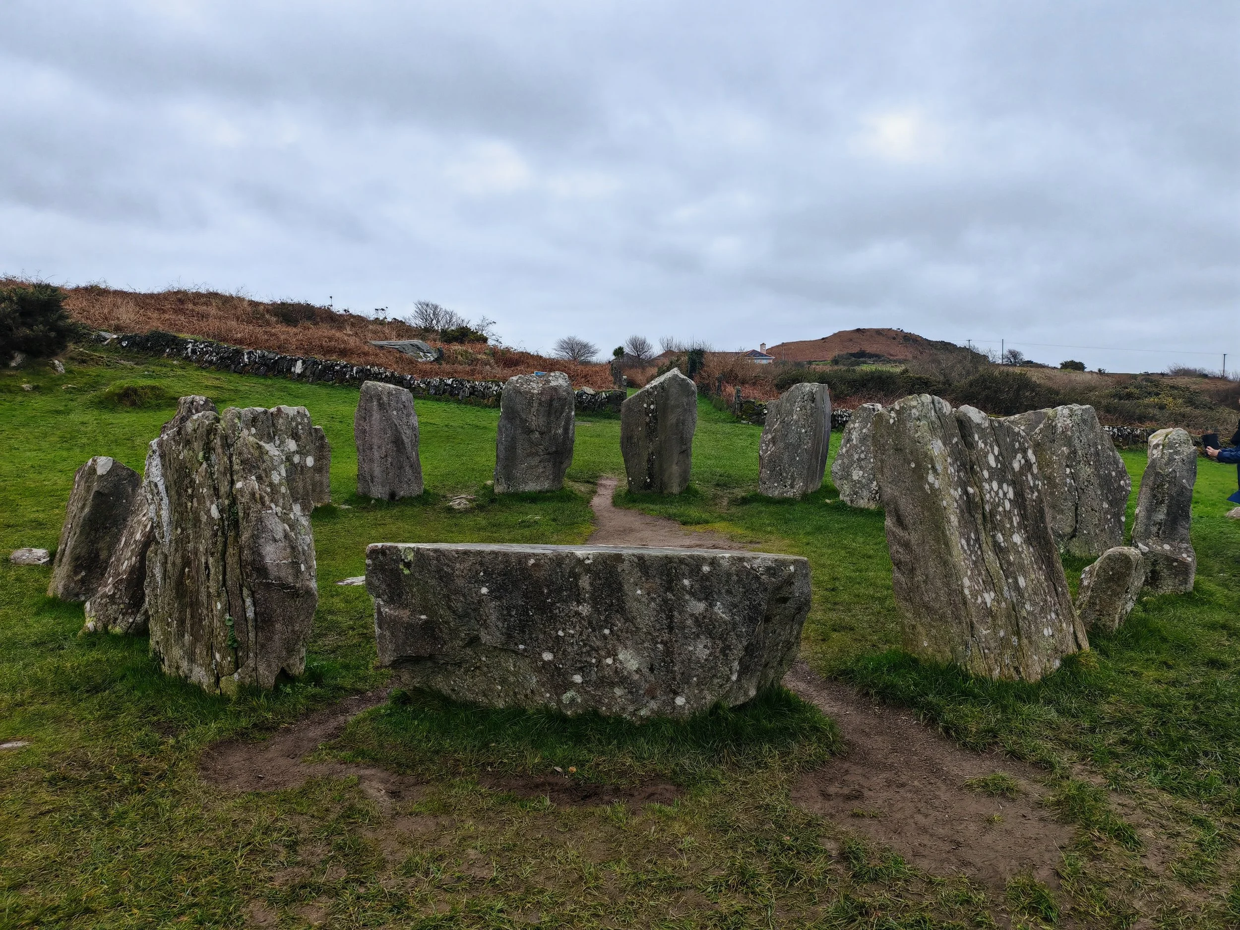 A prehistoric stone circle with large upright stones arranged in a ring on a grassy field under a cloudy sky, with a dirt path leading through the circle.