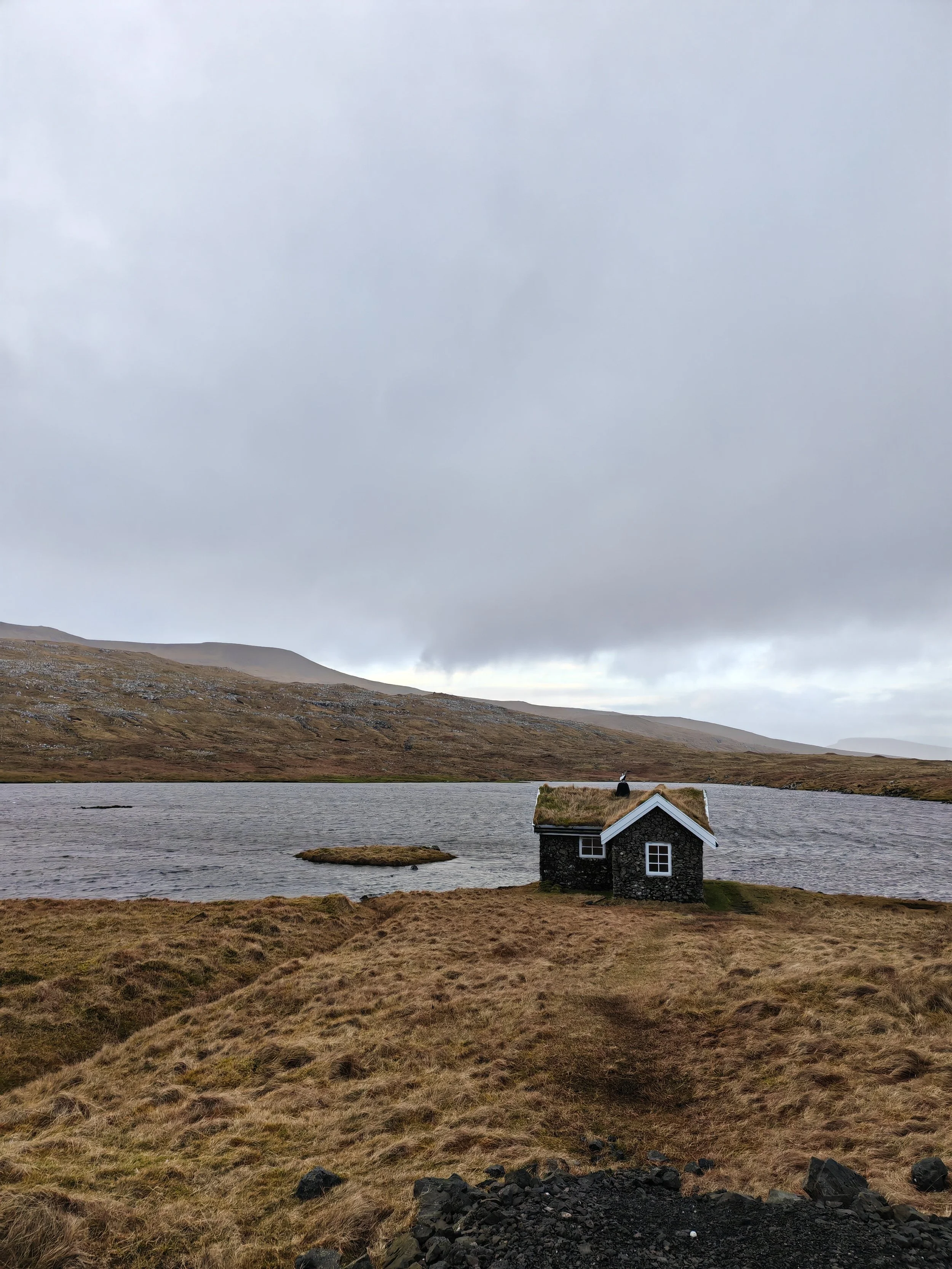A small stone house with a grass-covered roof sits by a lake in a mountain landscape under a cloudy sky.
