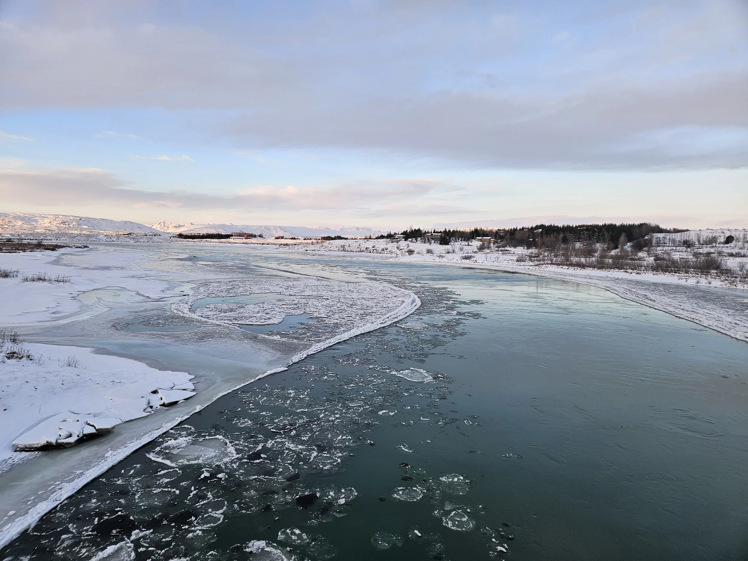 Snow-covered landscape with a partially frozen river, distant snow-capped mountains, and a partly cloudy sky.