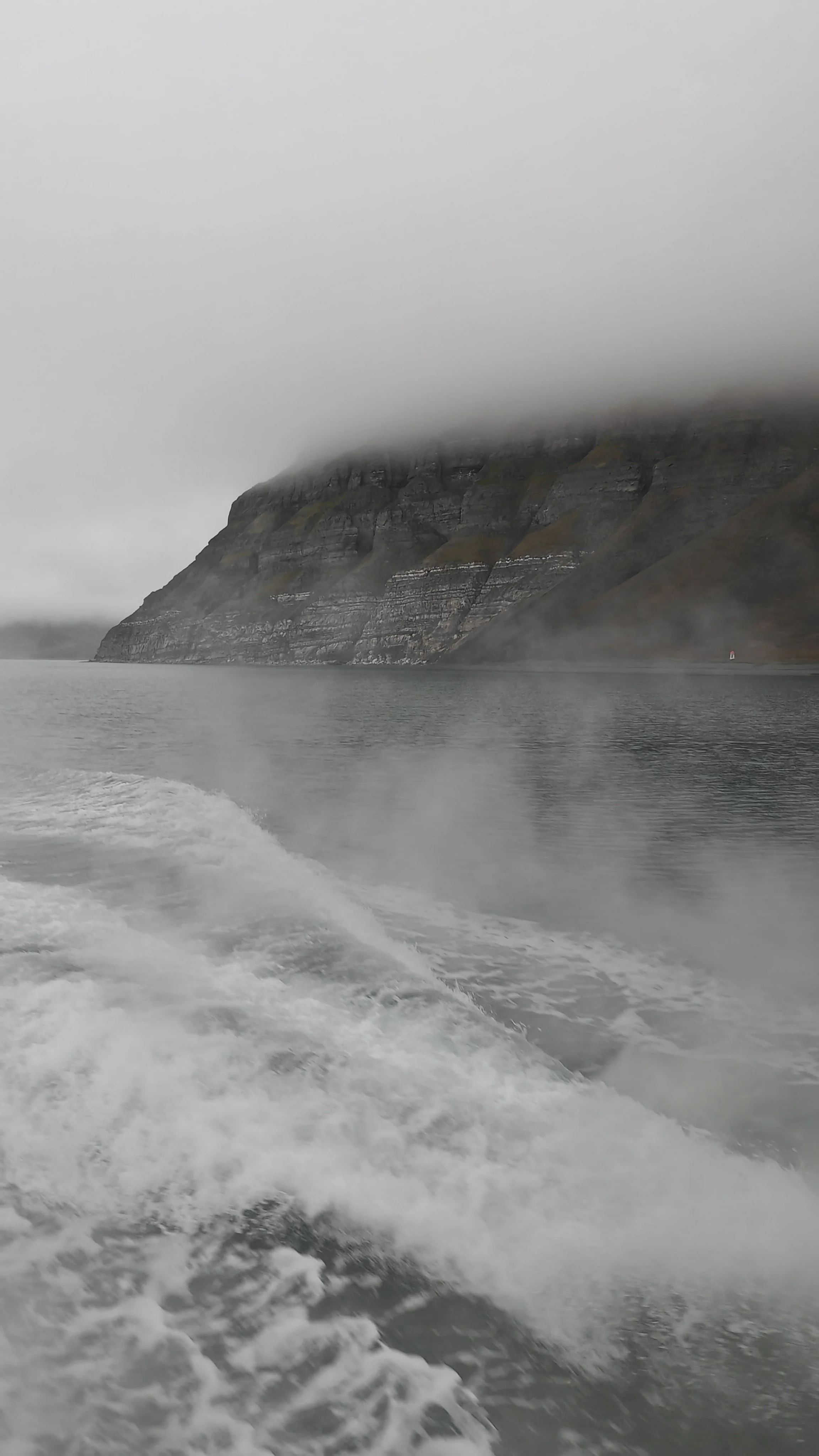 A large, fog-covered mountain with steep, layered rock faces along a body of water, viewed from a boat in motion.