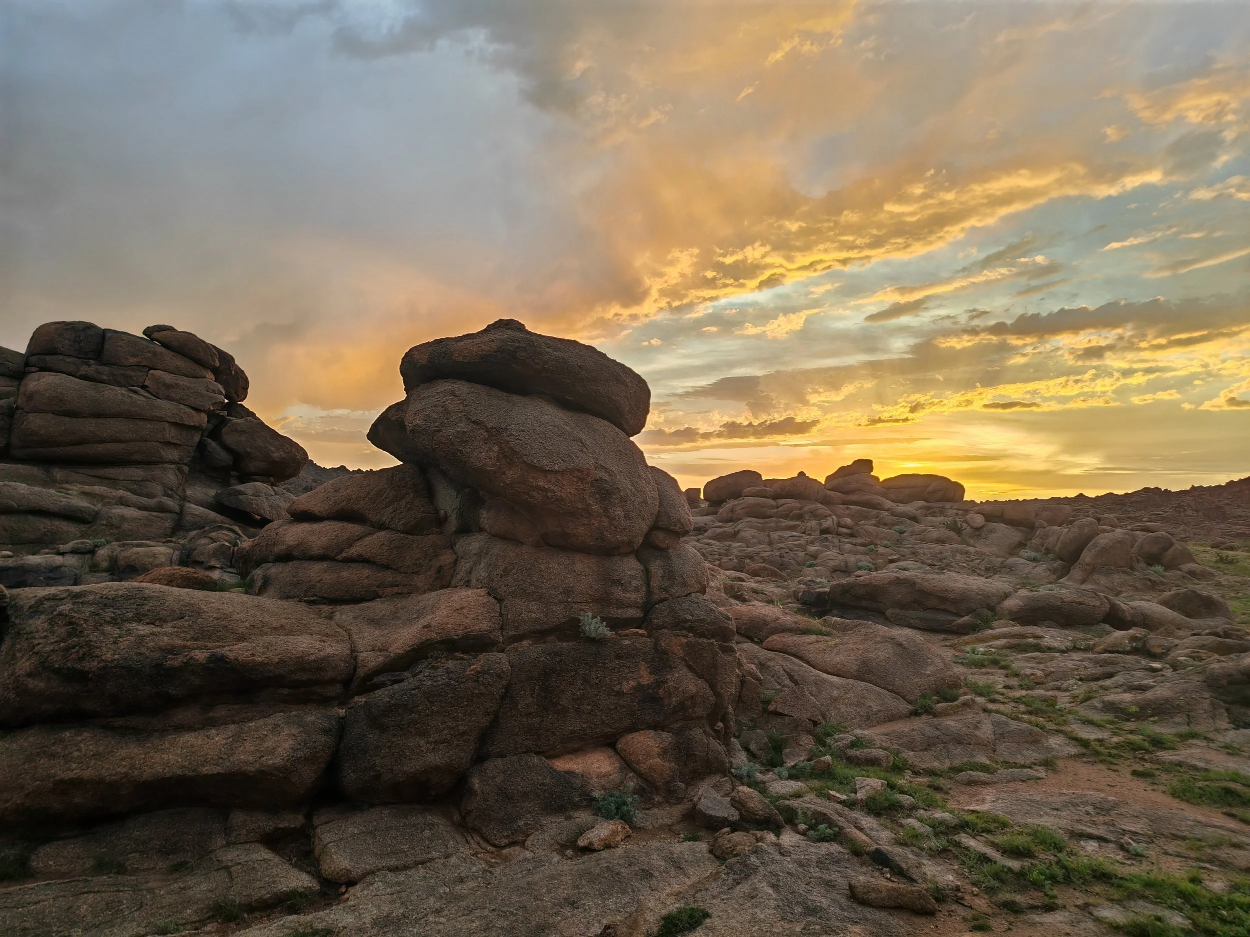 Large rocks and boulders scattered across a desert landscape at sunset, with a colorful sky of clouds and golden hues