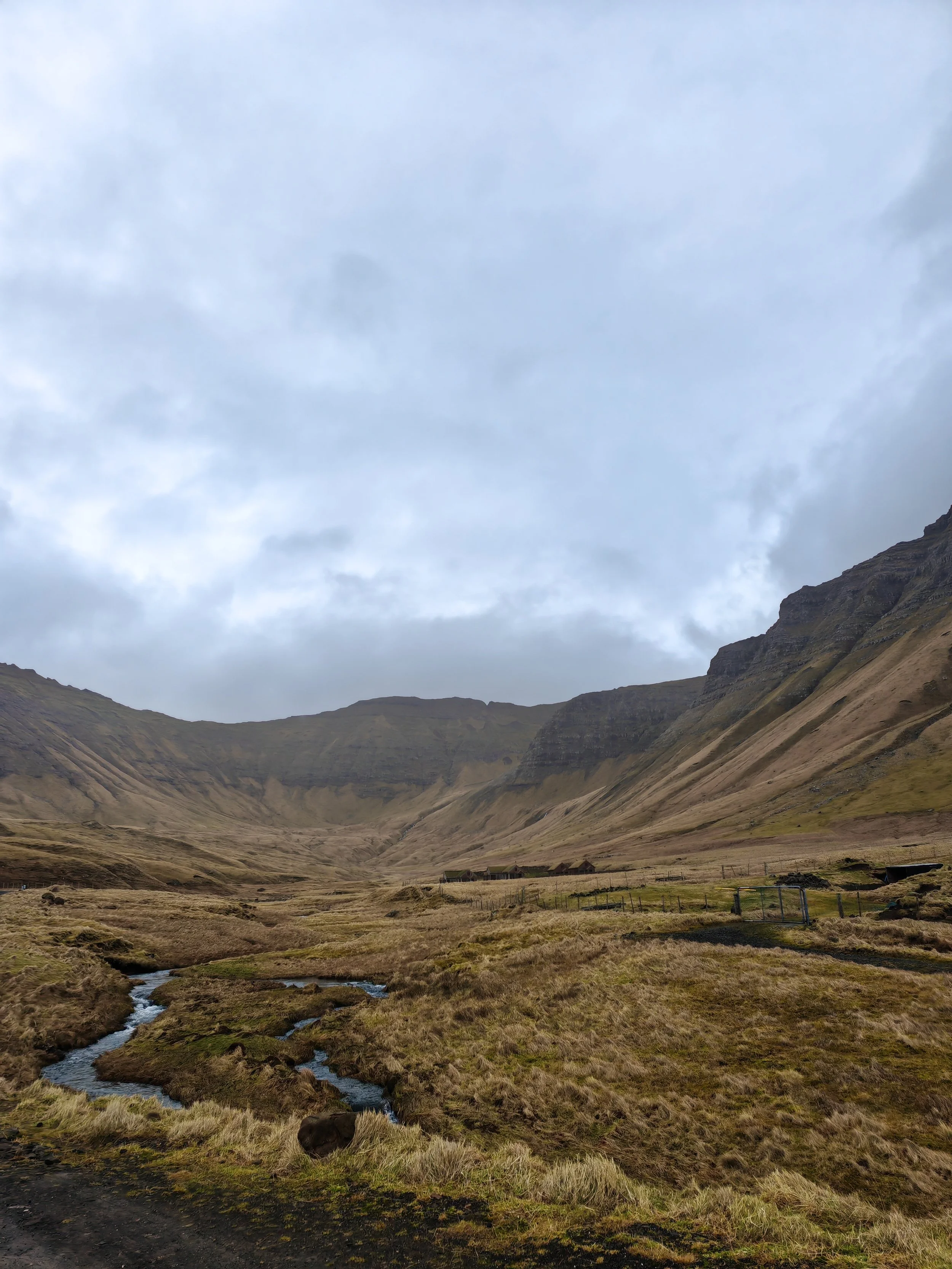 A vast mountainous landscape with a small stream flowing through grassy fields, mountains in the distance under a cloudy sky.