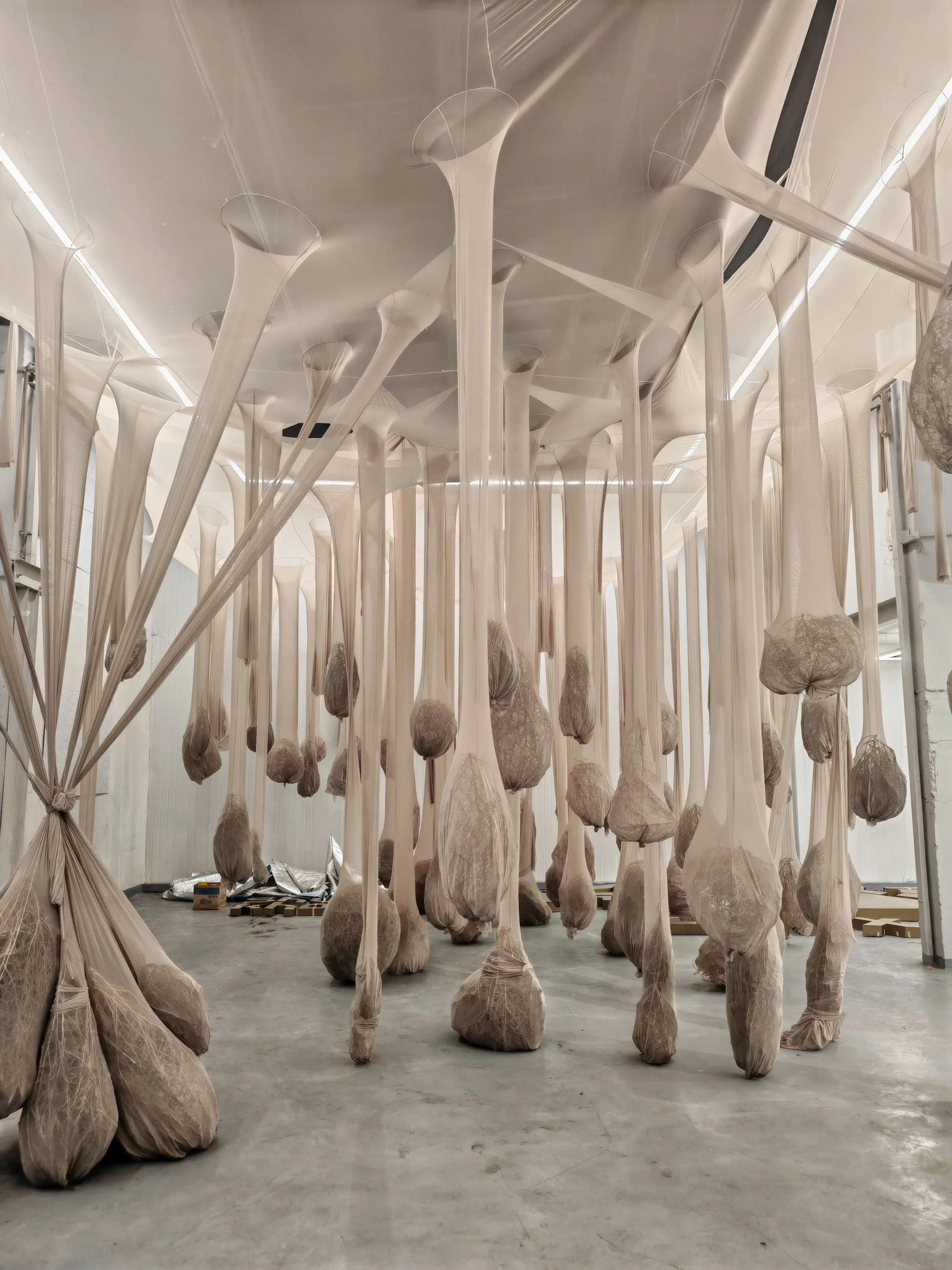 Art installation featuring numerous tall, elongated shapes covered with fabric, hanging from ceiling and standing on floor in a large open space.