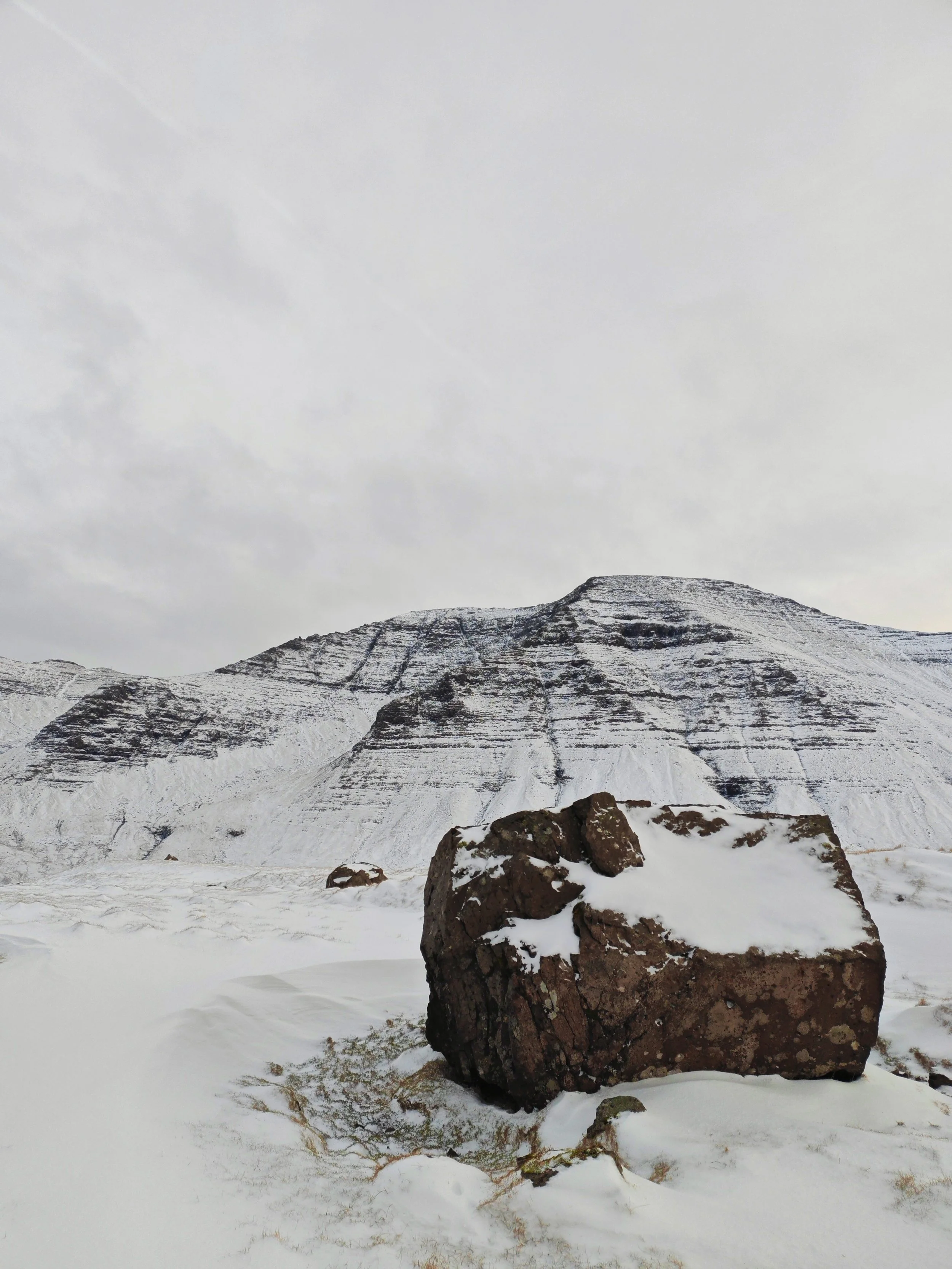 A snow-covered mountain landscape with large rocks in the foreground and a cloudy sky.