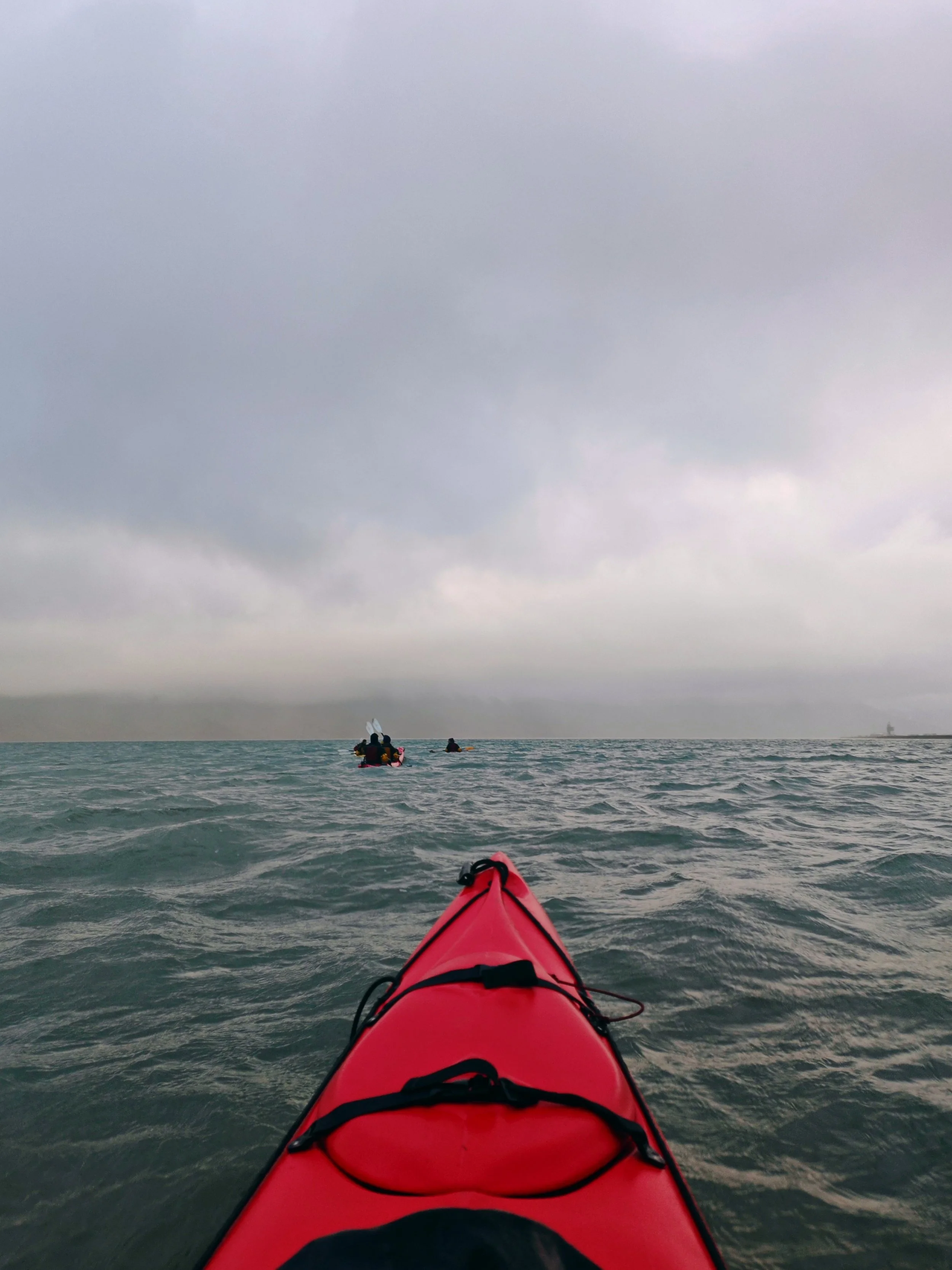 View from a kayak on the ocean during overcast weather, with other kayakers in the distance and a ship on the horizon.
