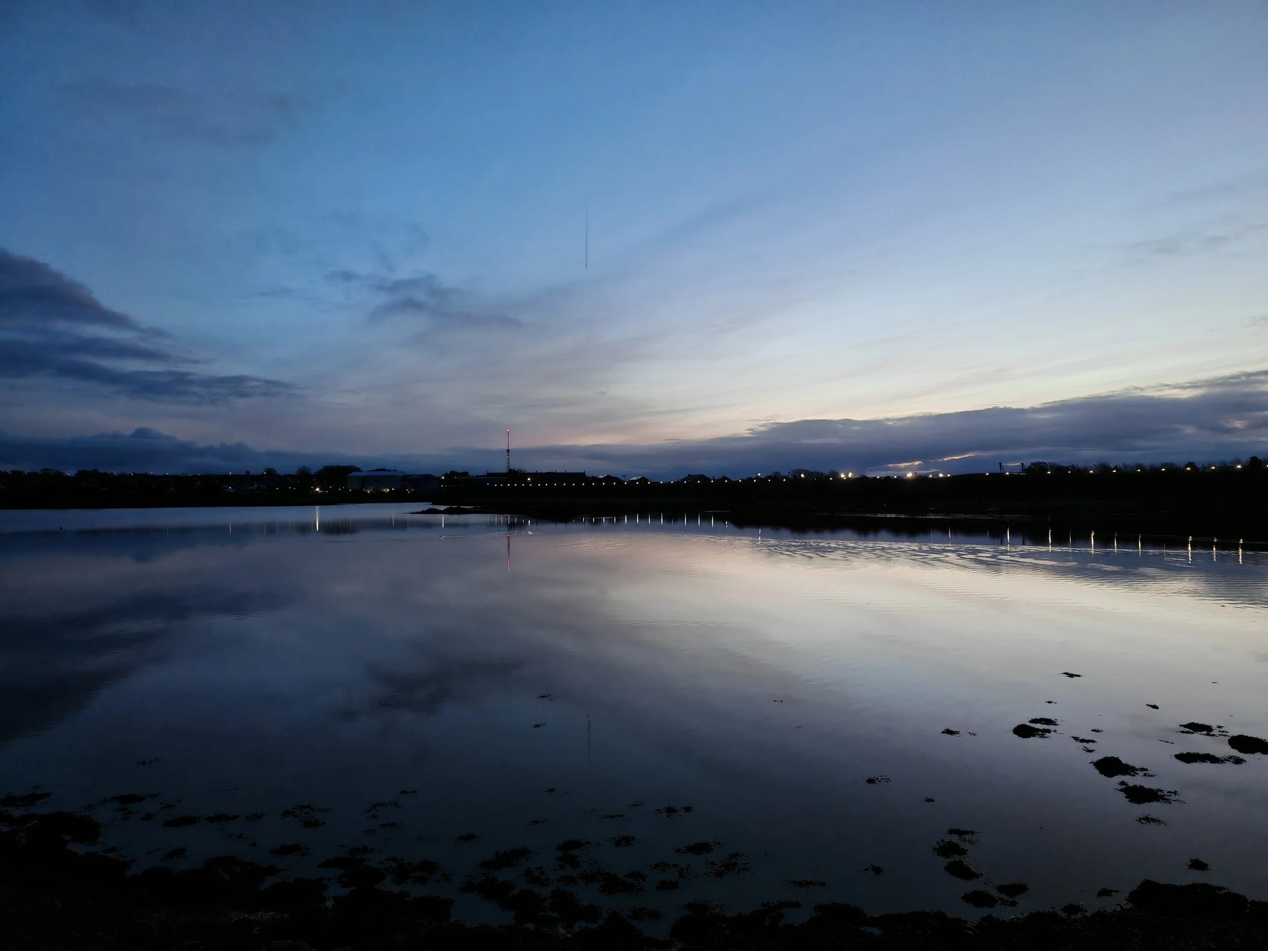 A lake or river at dusk with a calm, reflective surface, a cloudy sky with streaks of blue and gray, and a distant shoreline with small buildings and trees.