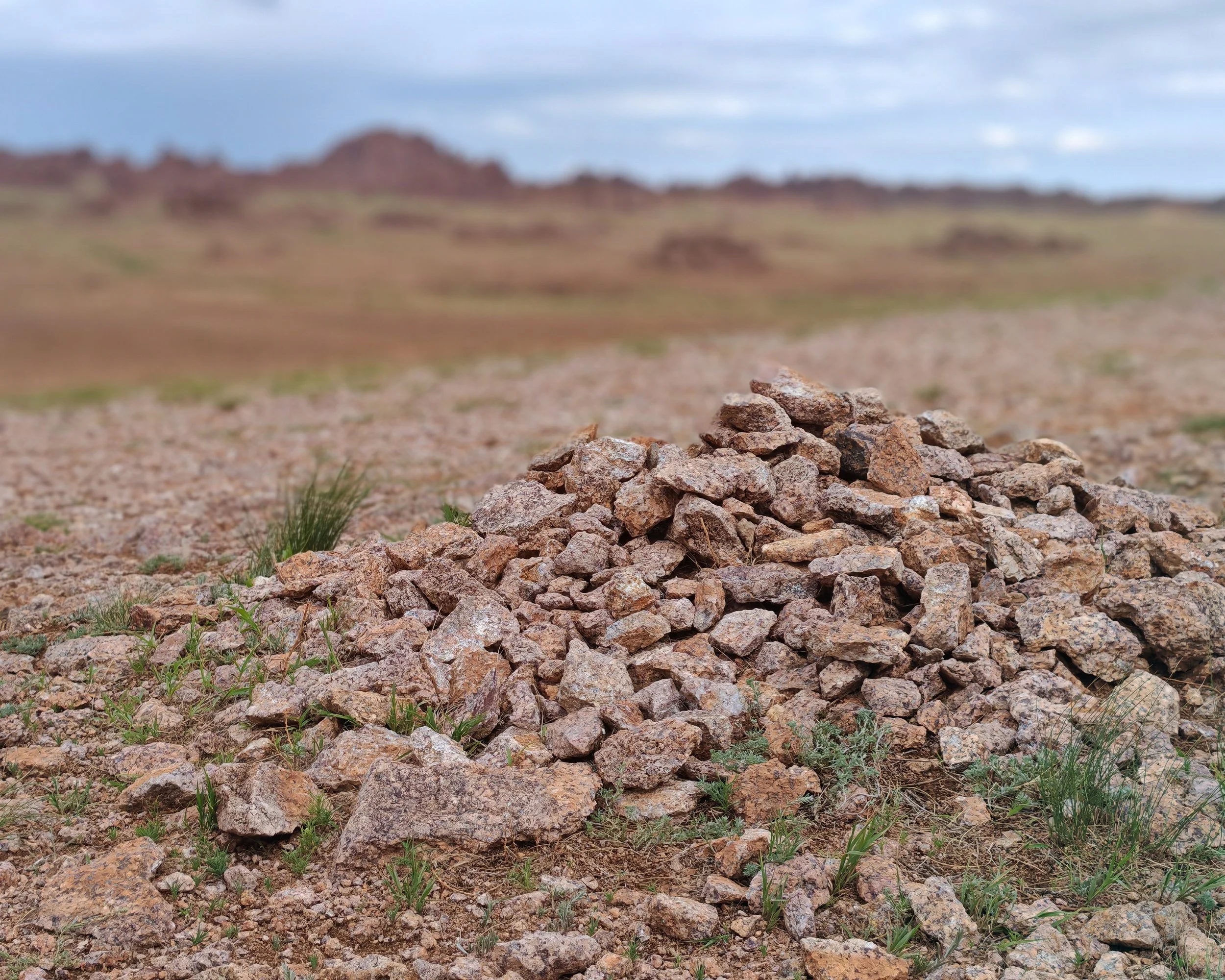 A pile of rocks and gravel in a desert landscape with distant rocky hills or mountains under a cloudy sky.