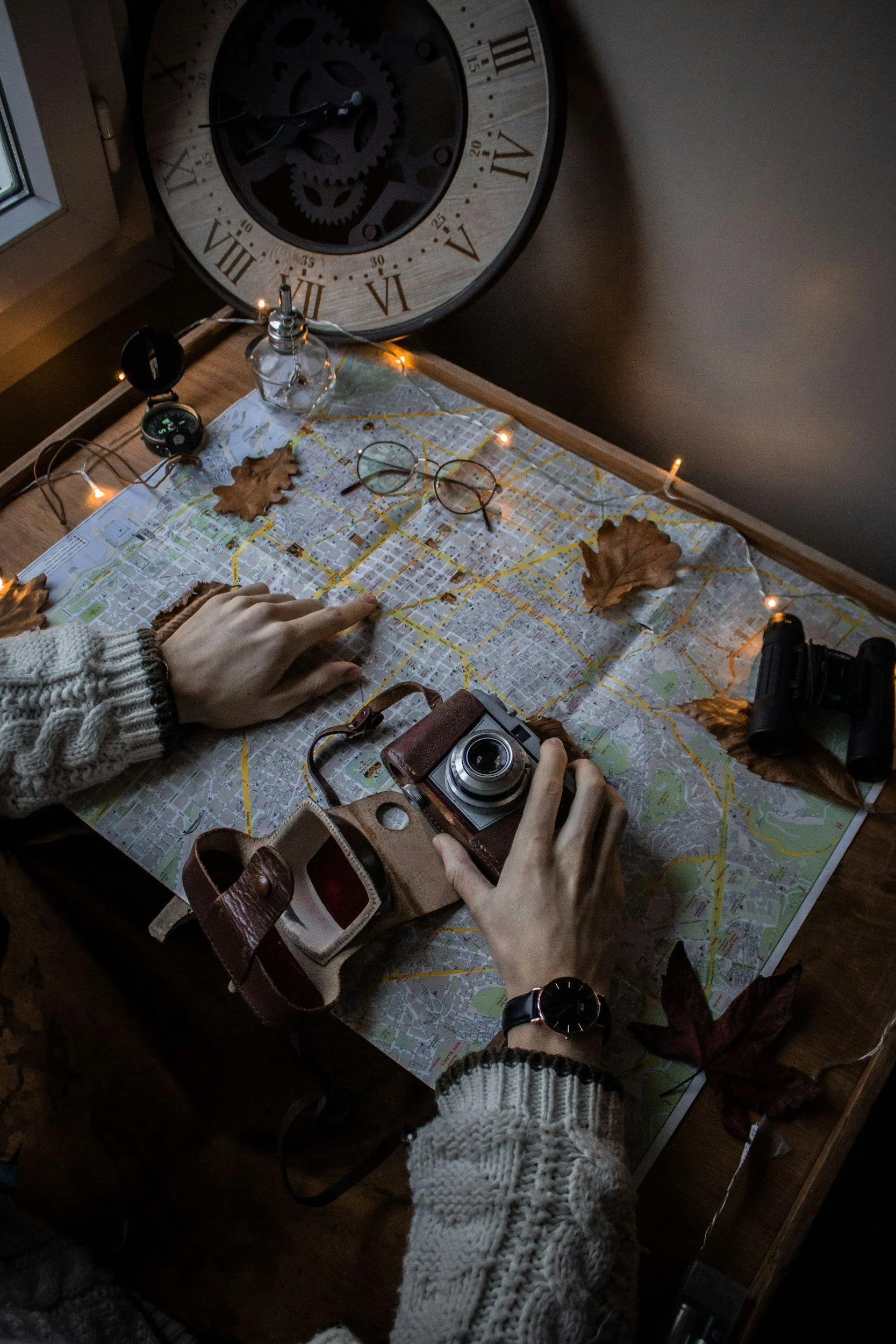 A table with a city map, a vintage camera, a leather purse, and a pair of glasses. There are dried leaves on the table and string lights. A person's hand is pointing at the map, and other hands are nearby.