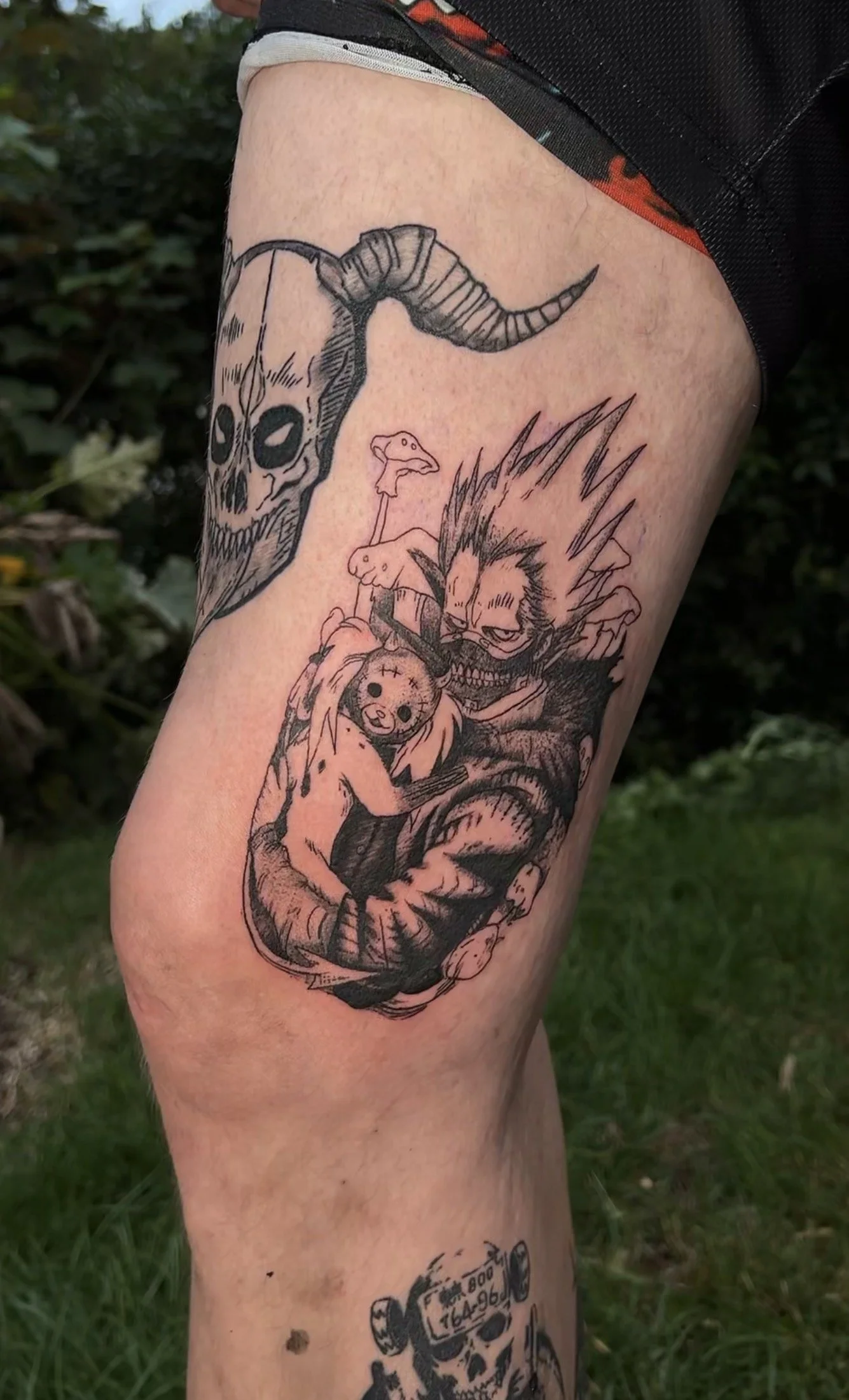 Tattoo auf dem Oberarm mit einem Dämonen- und Tiermotiv in Schwarzweiß, das einen Wolfskopf mit Hörnern, einen lachenden Dämon mit spitzen Zähnen und ein kleines, flauschiges Tier zeigt.
