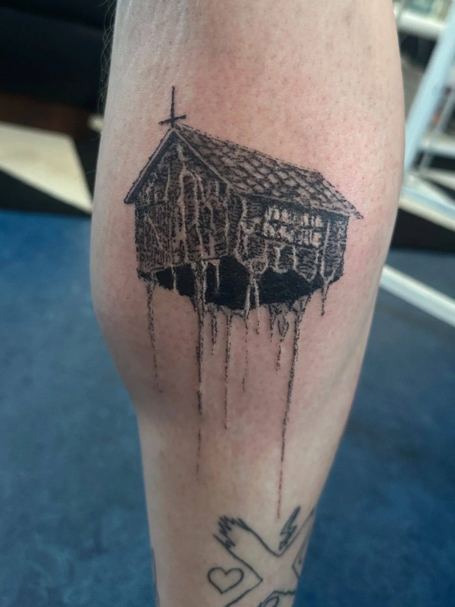 Tattoo eines Hauses mit schrägem Dach und Fenstern auf einem Bein, in Schwarz und Grau. Es gibt auch andere Tattoos auf dem Bein.