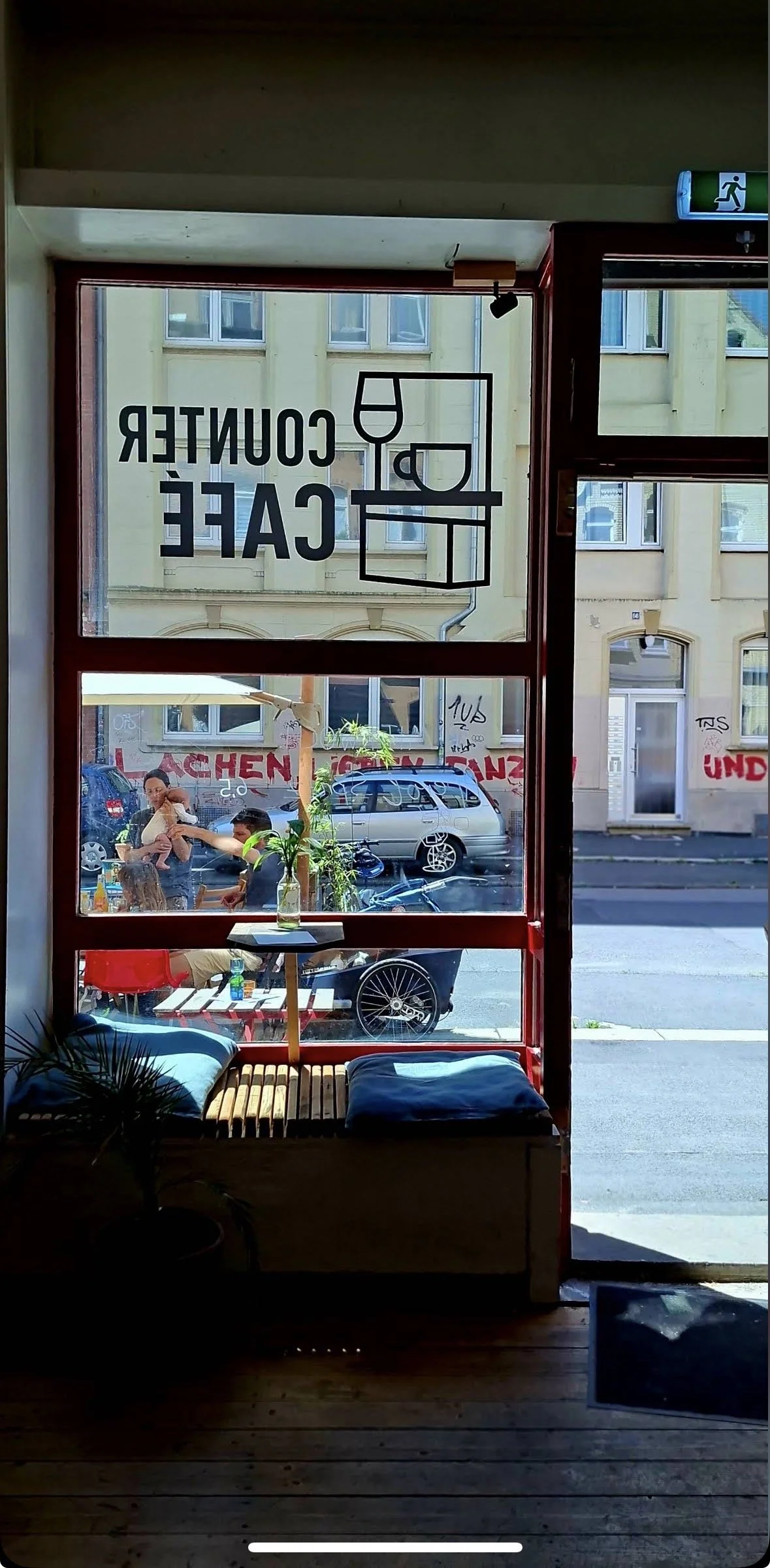 Blick durch eine Fensterscheibe in ein Café. Im Inneren sind zwei Sitzbänke mit Kissen, ein kleiner Tisch, auf dem eine Wasserflasche steht. Draußen sind Menschen, Autos und Graffiti an einer Wand sichtbar.
