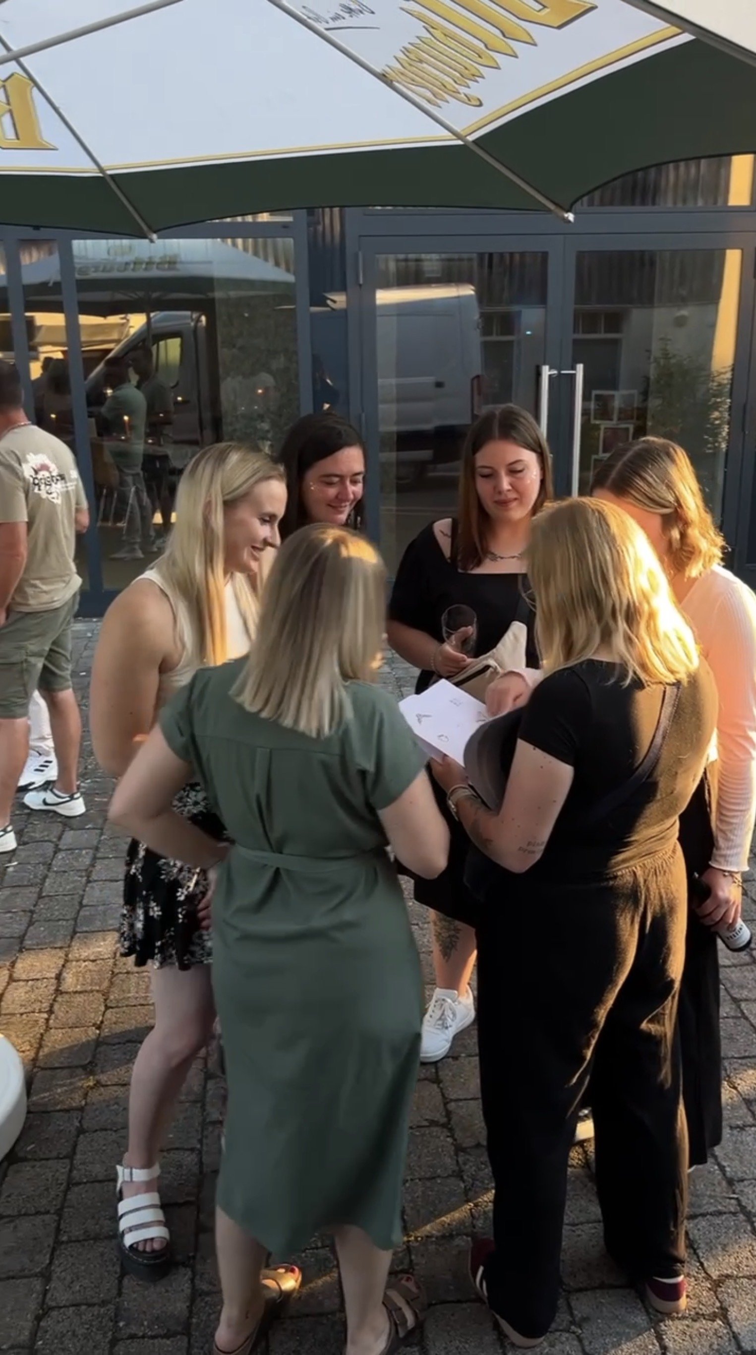 Eine Gruppe von Frauen steht im Freien, sie schauen auf ein Smartphone und lachen. Es ist Abend, und sie befinden sich vor einem Gebäude mit großen Fenstern, unter einem Sonnenschirm.