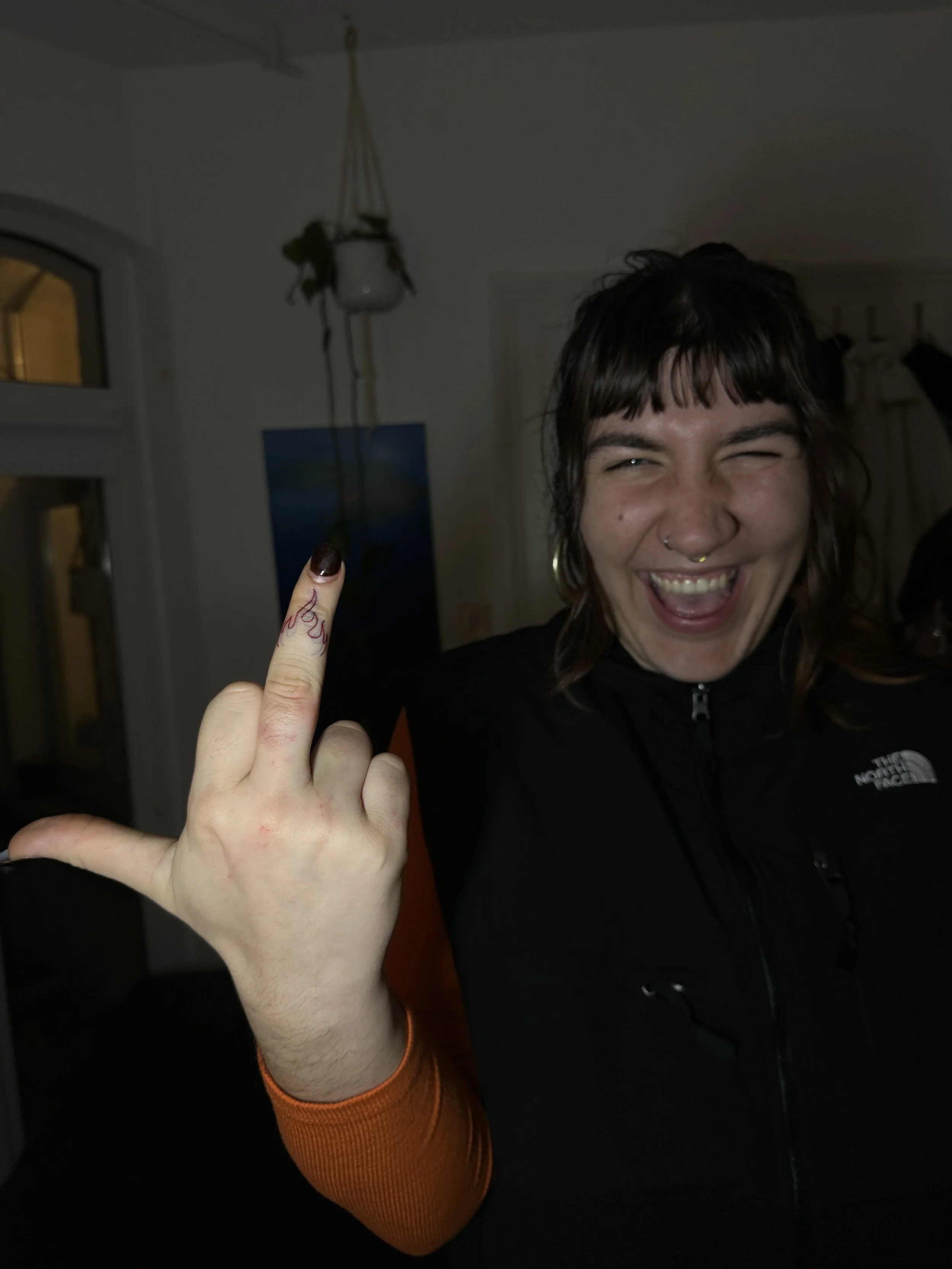Eine lachende Frau mit dunklen Haaren und Piercings zeigt auf ihren Finger, der mit roten Markierungen und Tattoos verziert ist, in einem dunklen Raum mit Fenster und Pflanze im Hintergrund.