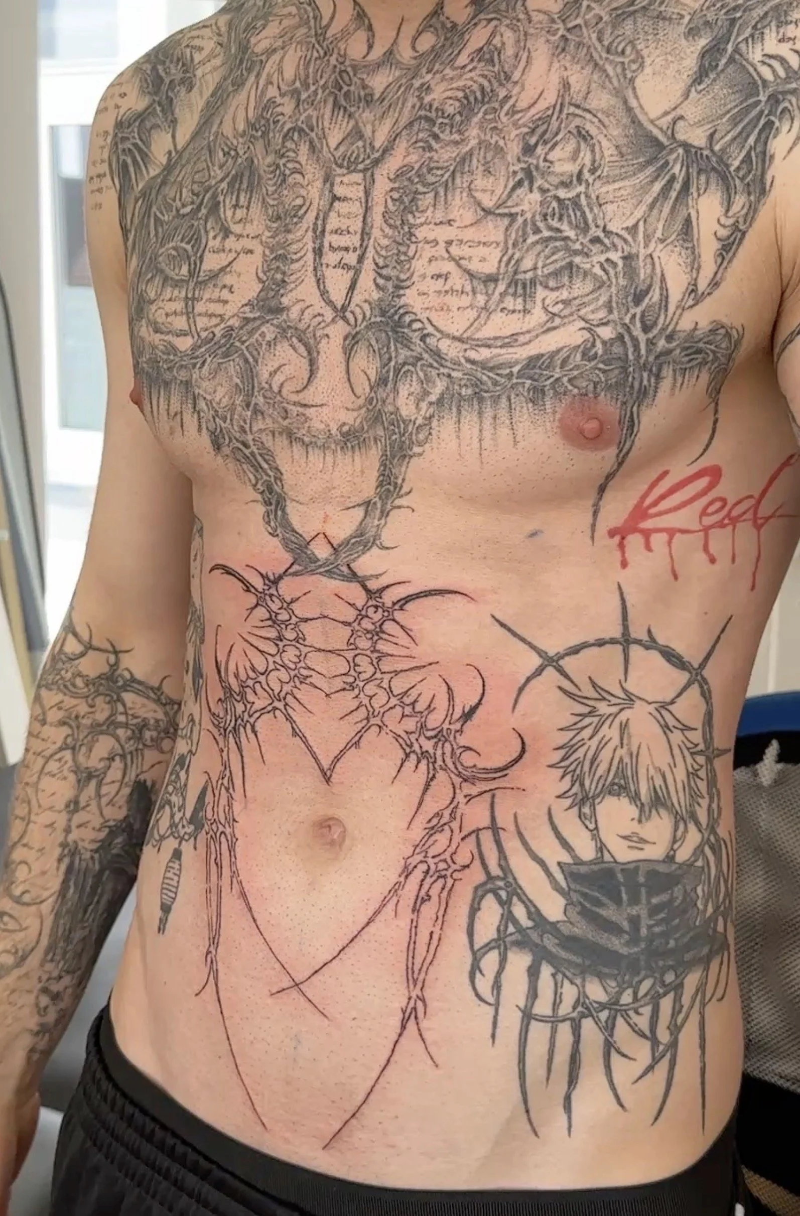 Nahaufnahme eines männlichen Oberkörpers mit zahlreichen schwarzen Tattoo-Designs, darunter eine große zentrale Figur, Sonnen- und Mondsymbole sowie ein Comic-ähnliches Gesicht, alles auf nackter Haut.