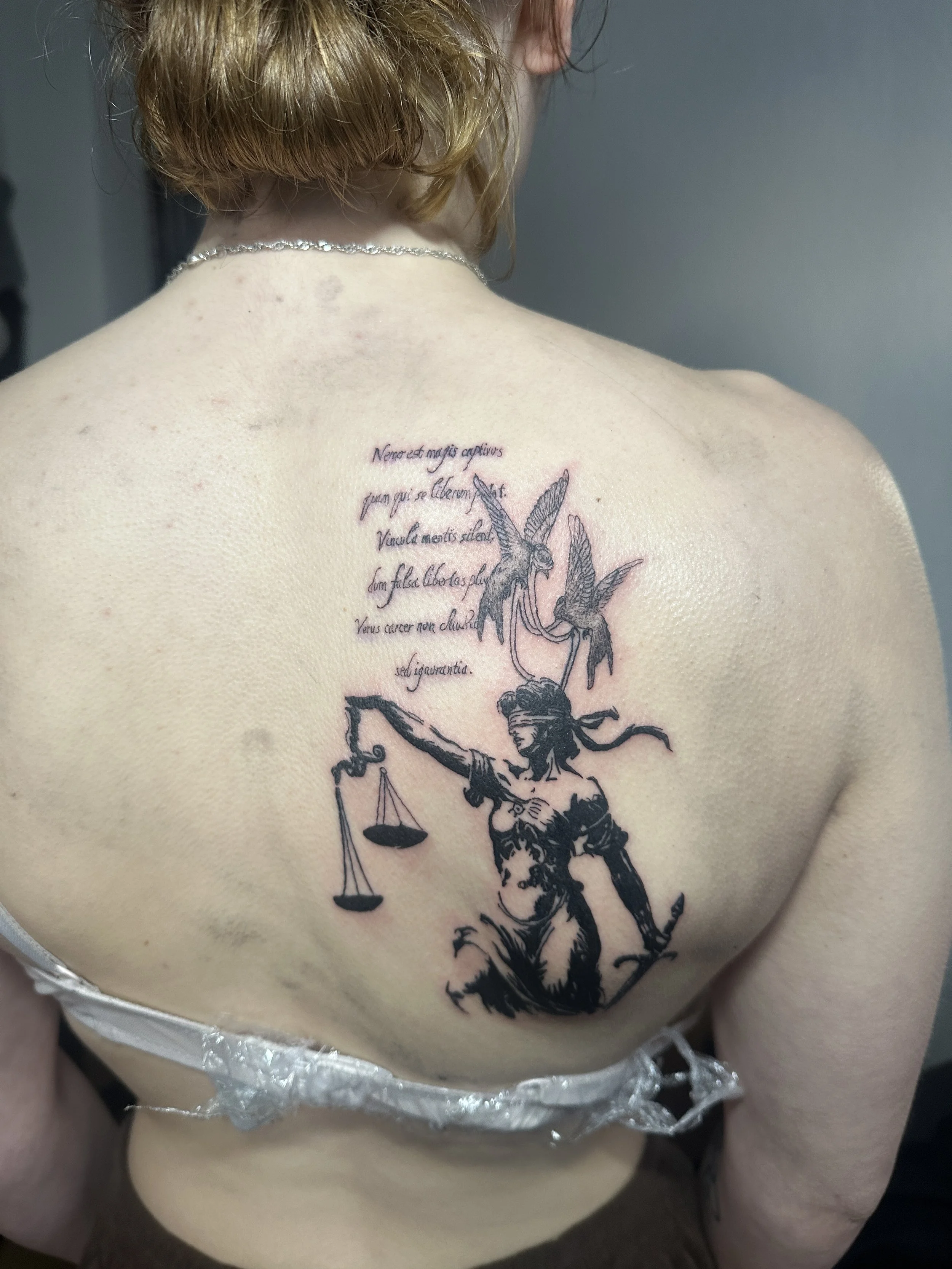 Tattoo auf dem Rücken einer Person, zeigt die Statue der Gerechtigkeit mit Waage, Schwert und verbundenen Augen, begleitet von lateinischem Text.