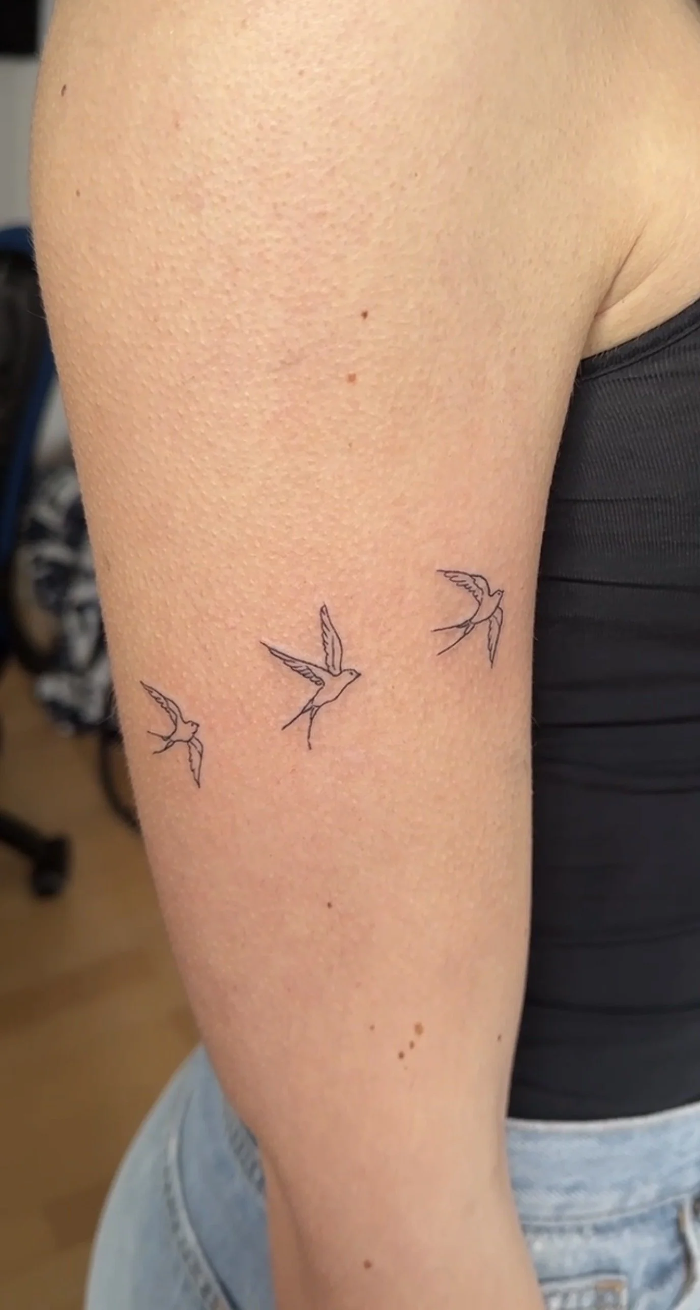 Drei kleine, einfache schwarze Tattoos von fliegenden Vögeln auf dem Oberarm einer Person.