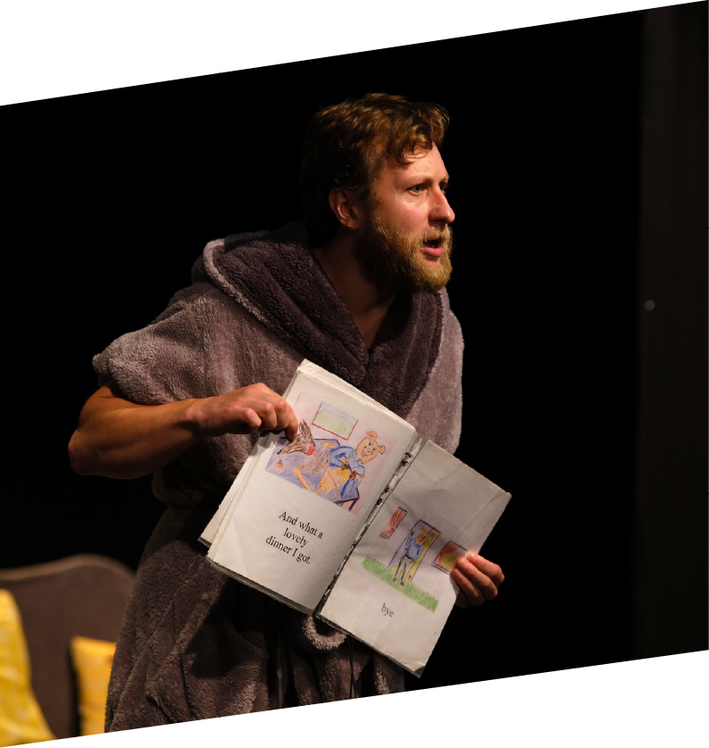 A man with a beard and curly hair, dressed in a thick towel, is performing on stage, holding an open children's book with colorful illustrations and text, in a dimly lit setting.