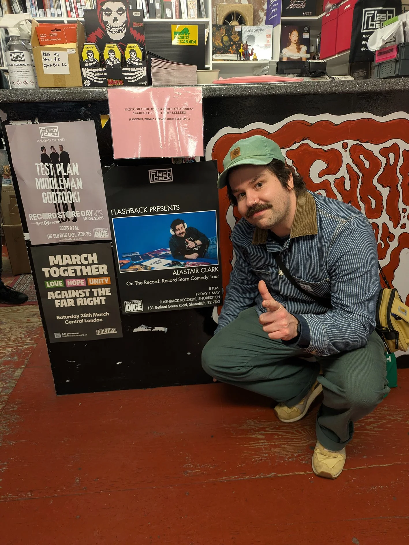 A man with a mustache, wearing a light green cap, striped jacket, and green pants, is crouching down and pointing at the camera, in front of a black counter with posters for music events and various signs, in an indoor setting with red wooden floor and shelves with books and items in the background.
