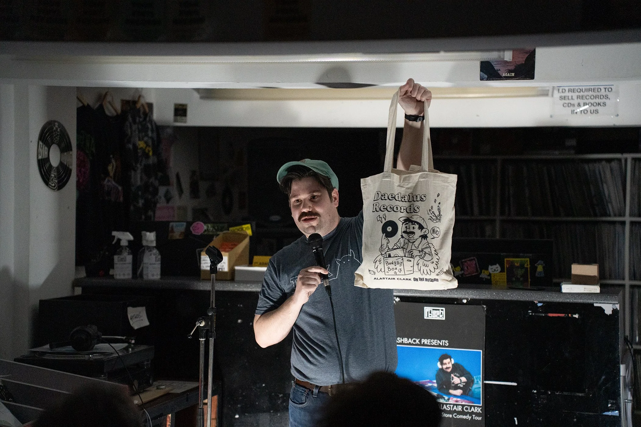 A man holding a tote bag with cartoon illustrations and text, speaking into a microphone at an indoor event, background showing posters, shelves with books, and a sign about selling records, CDs, and books.