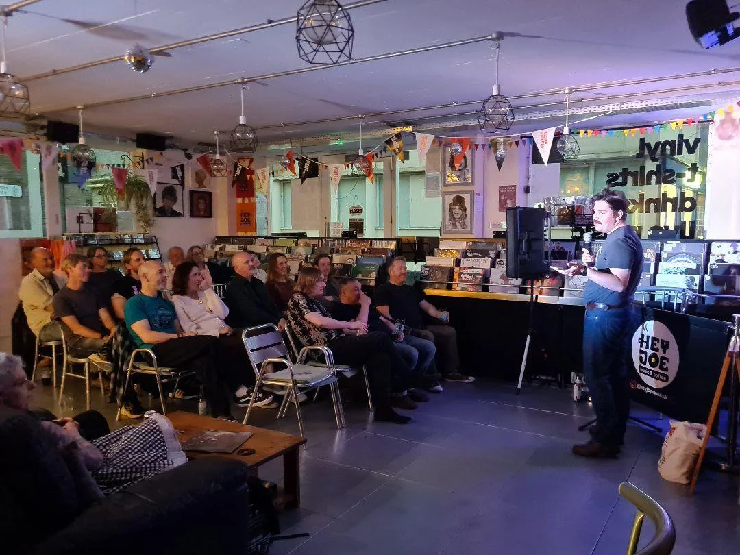 A man performs stand-up comedy for an audience in a cozy venue decorated with colorful bunting, photographs, and vinyl records.