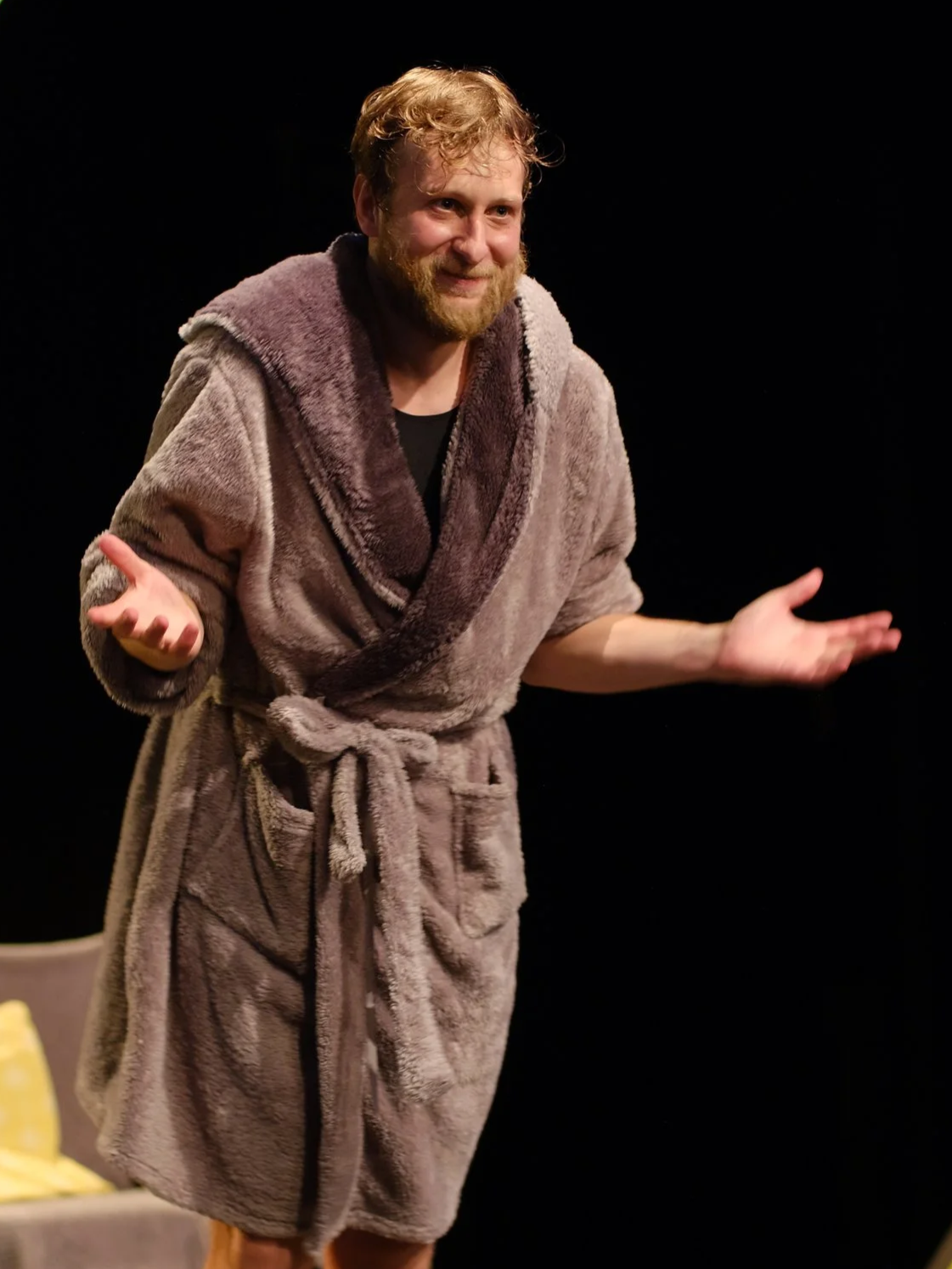 Man performing on stage in a brown plush bathrobe with a belt, smiling and gesturing with open hands, against a black background.
