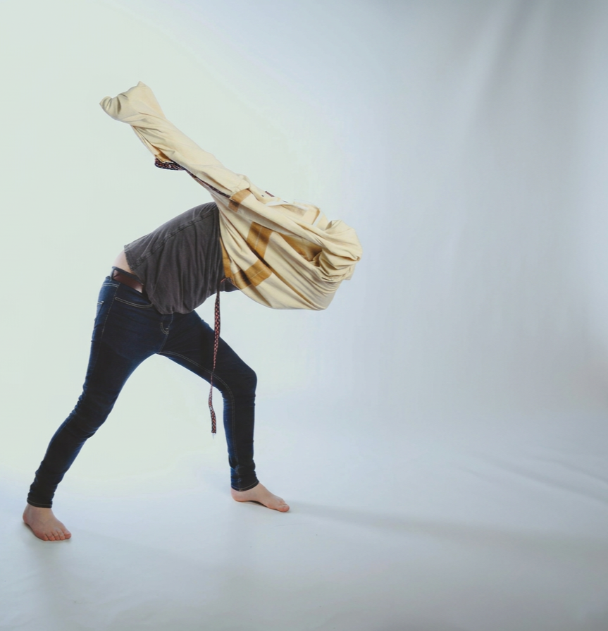 Person in casual clothing, barefoot, in a wide stance, throwing a jacket over their head in a studio with a plain background.