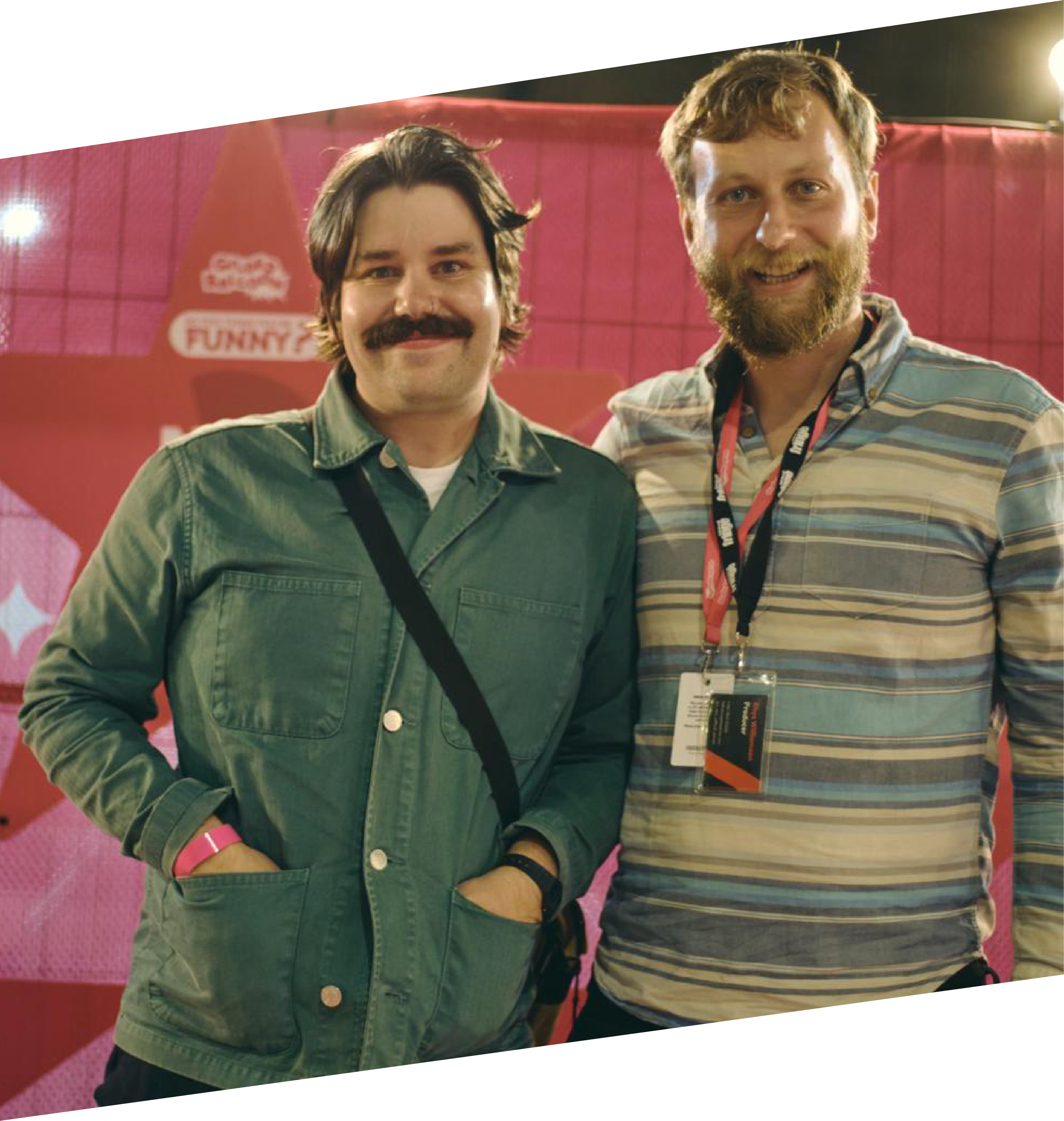 Two smiling men standing together at an indoor event with a pink background and a sign that reads 'Funny'.