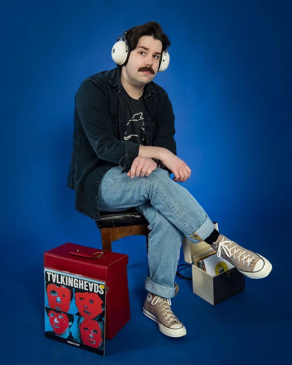 A man with dark hair and a mustache wearing headphones, a black denim jacket, black t-shirt, light blue jeans, and beige sneakers, sitting on a chair against a blue background, with a Talking Heads vinyl record and a small black basket nearby.