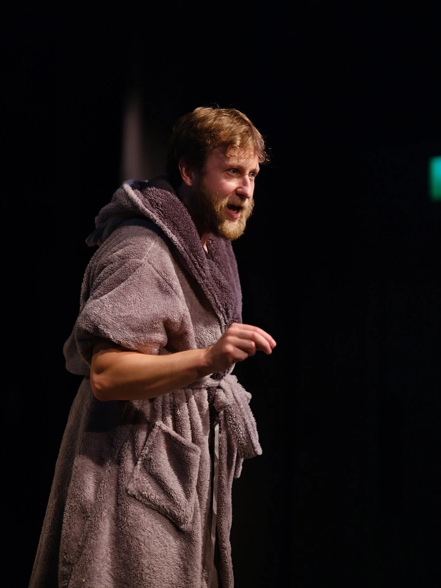 A man with a beard and light brown hair wearing a fuzzy, light purple bathrobe, appears to be speaking or explaining something on a stage or in a dark room with a black background.