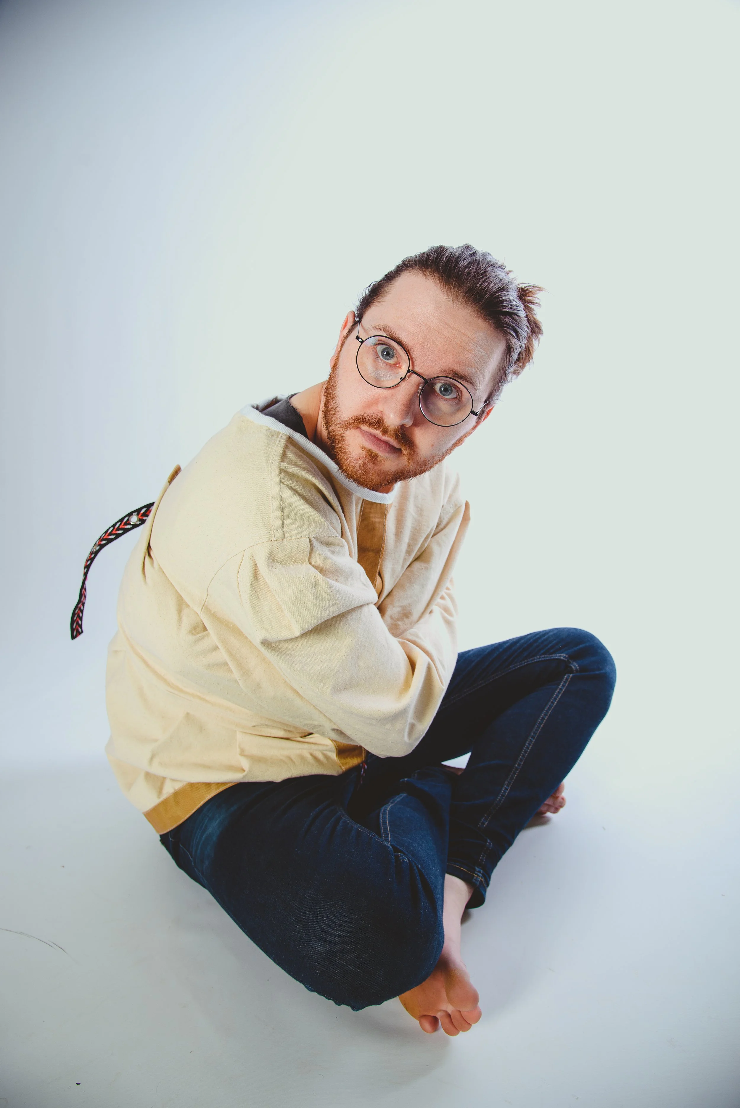 A man with glasses, a beard, and short hair, wearing a beige hoodie and dark jeans, sitting cross-legged on the floor against a plain light-colored background, looking directly at the camera.