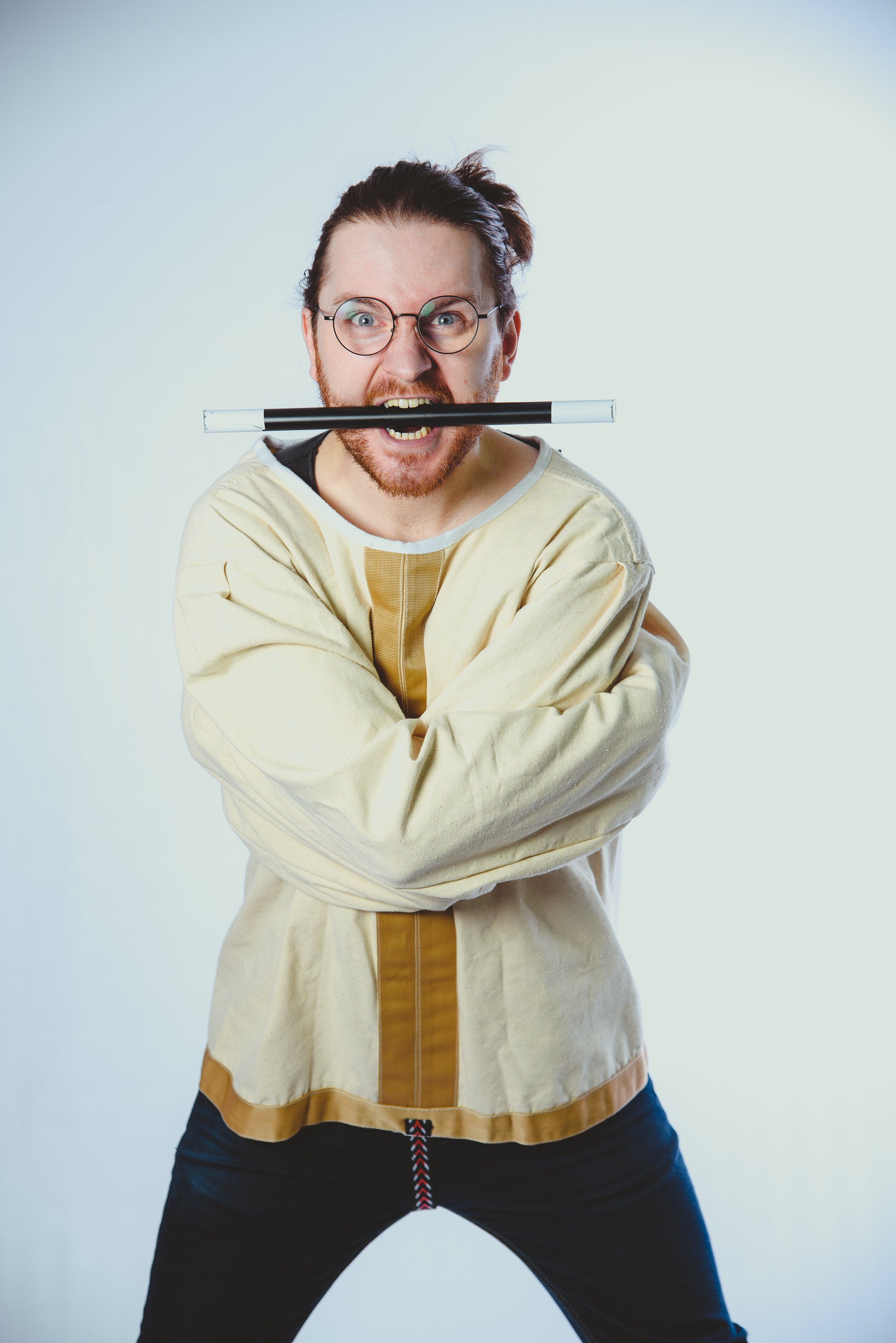 A man with glasses, red hair, and a beard with a pen in his mouth, crossing his arms and standing with legs apart, wearing a beige and brown long-sleeve shirt, against a plain light background.