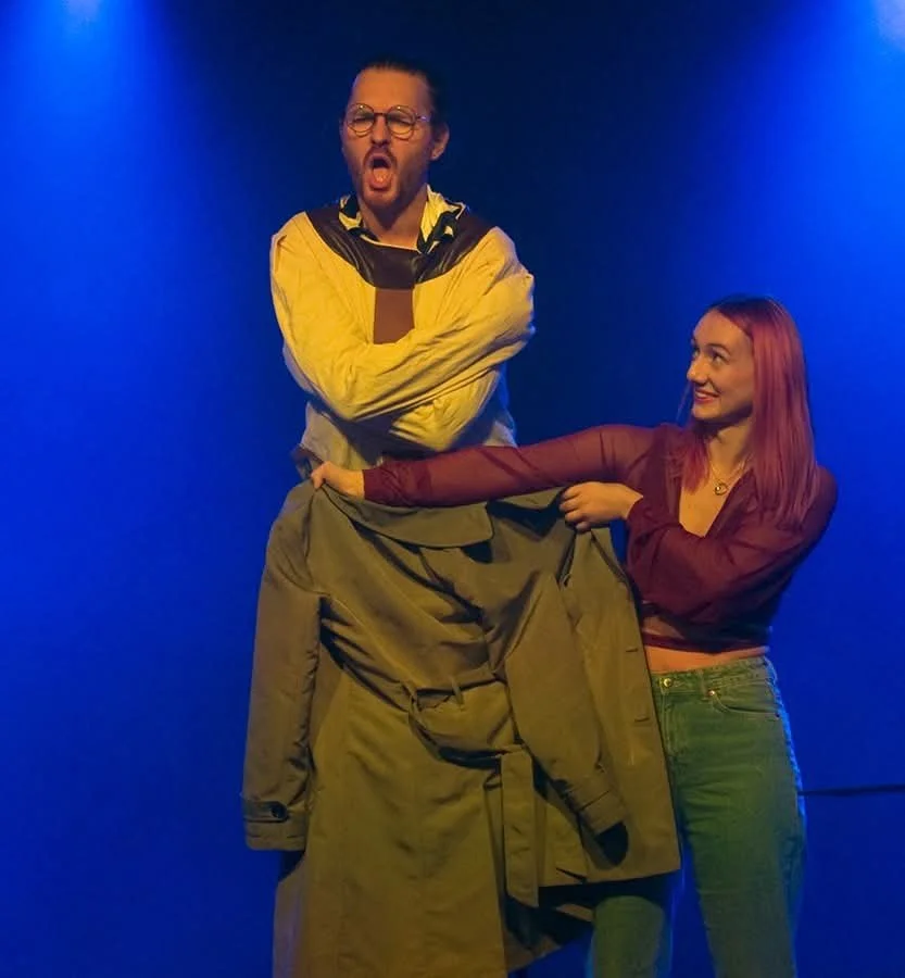 Two people, a man with glasses, a beard, and long hair, and a woman with long pink hair, stand against a blue background. The man appears to be surprised or shocked, while the woman is smiling and pulling at the man's coat.