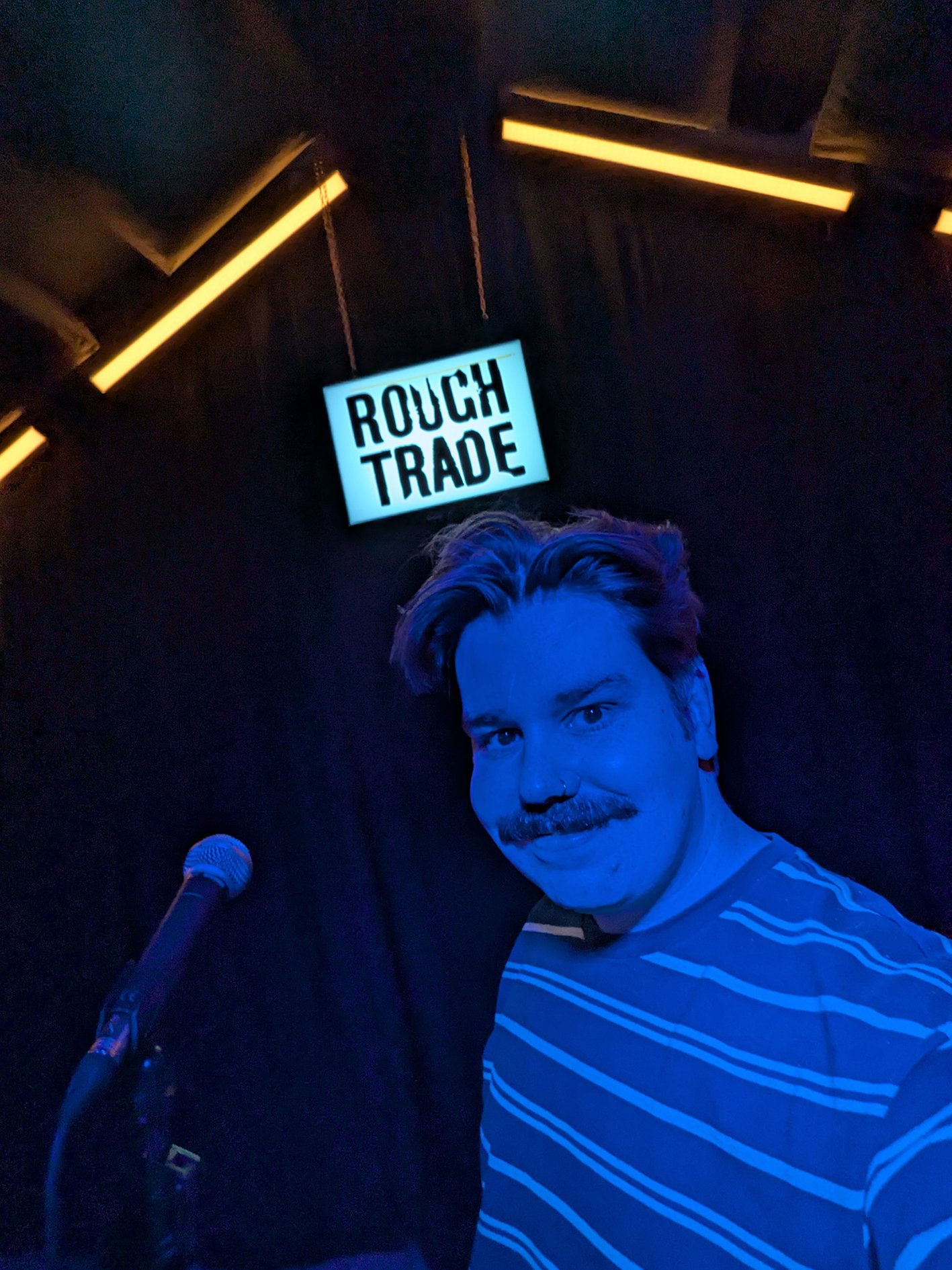 A person with a brown mustache and wavy hair, wearing a striped shirt, taking a selfie in a dark room. There is a sign that reads 'ROUGH TRADE' in the background and a microphone in the foreground.