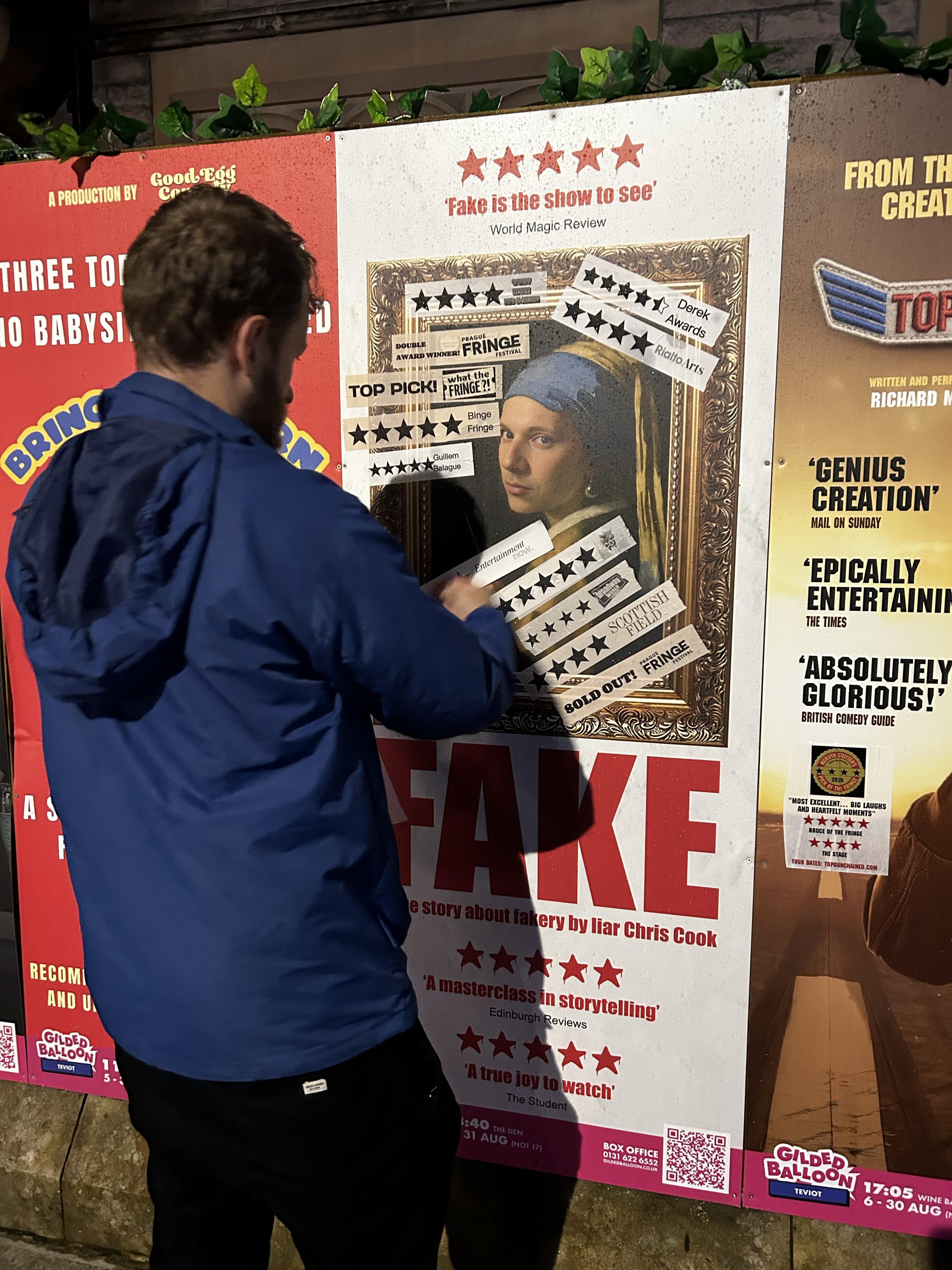 Man with dark curly hair wearing a blue jacket reading a poster on a wall.