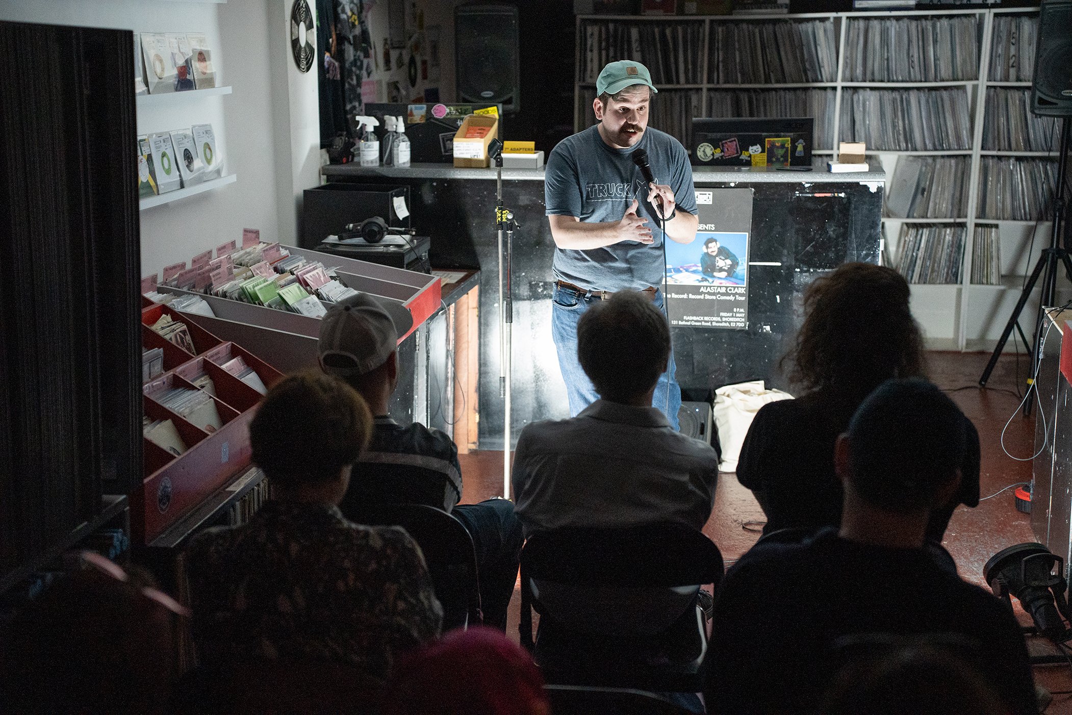 A man with a mustache and beard, wearing a green cap and a T-shirt, stands in front of an audience, holding a microphone and speaking. Behind him, there are shelves filled with records and a poster for Alastair Clark's comedy tour. The setting appears to be a record store or similar venue with merchandise on display and people seated watching him.