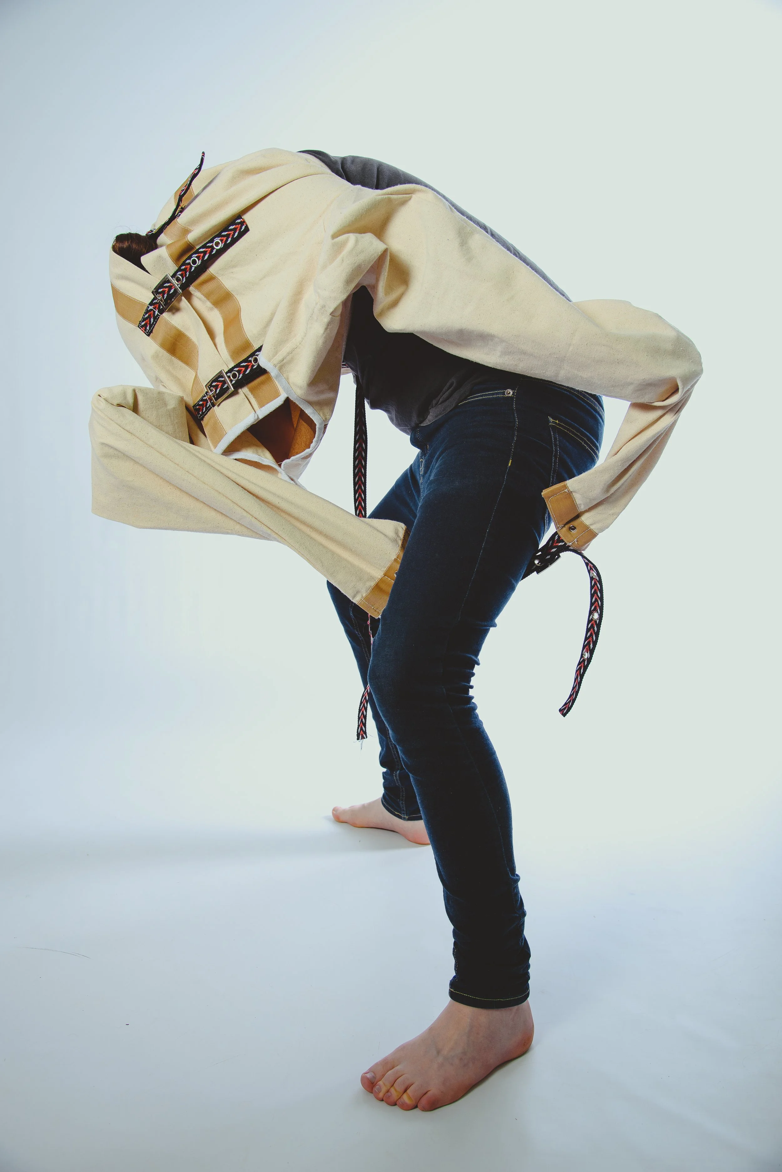 A person with long hair in a braid is standing barefoot on a white background, wearing dark jeans and a beige hoodie, with their body bent forward and arms extended, as if practicing a yoga pose.