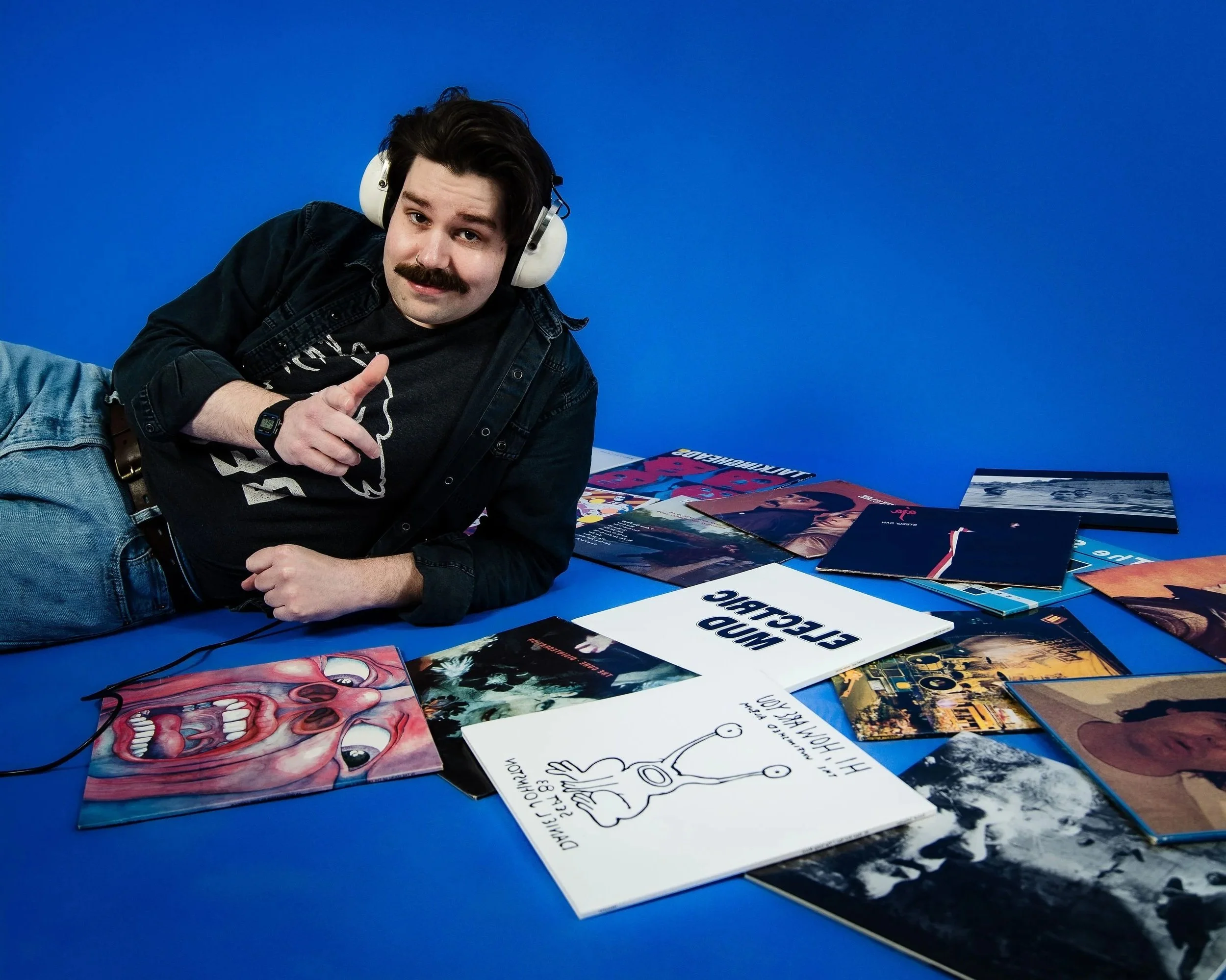 A man with dark hair, a mustache, and a goatee lying on a blue surface, wearing headphones and a black denim jacket, giving a thumbs-up gesture. There are various colorful posters and flyers spread out on the surface next to him.