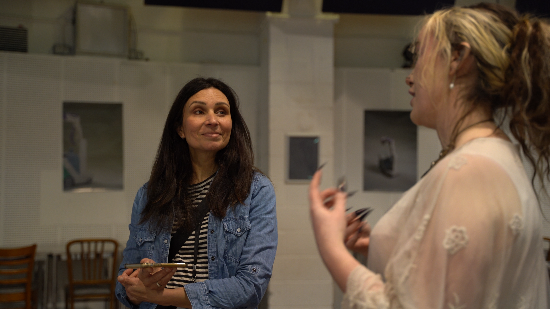 Two women engaged in conversation in a room with wooden chairs and white walls. One woman, with dark hair, wearing a denim jacket and striped shirt, has a slight smile and holds a smartphone. The other woman, with curly hair and glasses, gestures as she speaks.