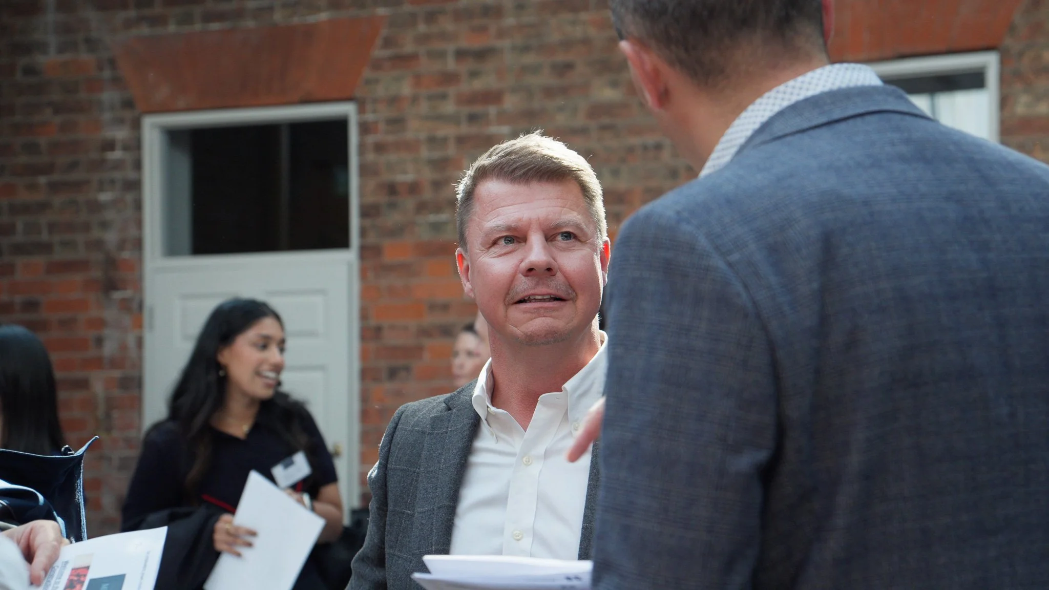 Two men are engaged in a conversation at an outdoor event, with one facing the camera and the other partially visible. Several women are in the background, smiling and holding papers. The setting features a brick wall and a window.