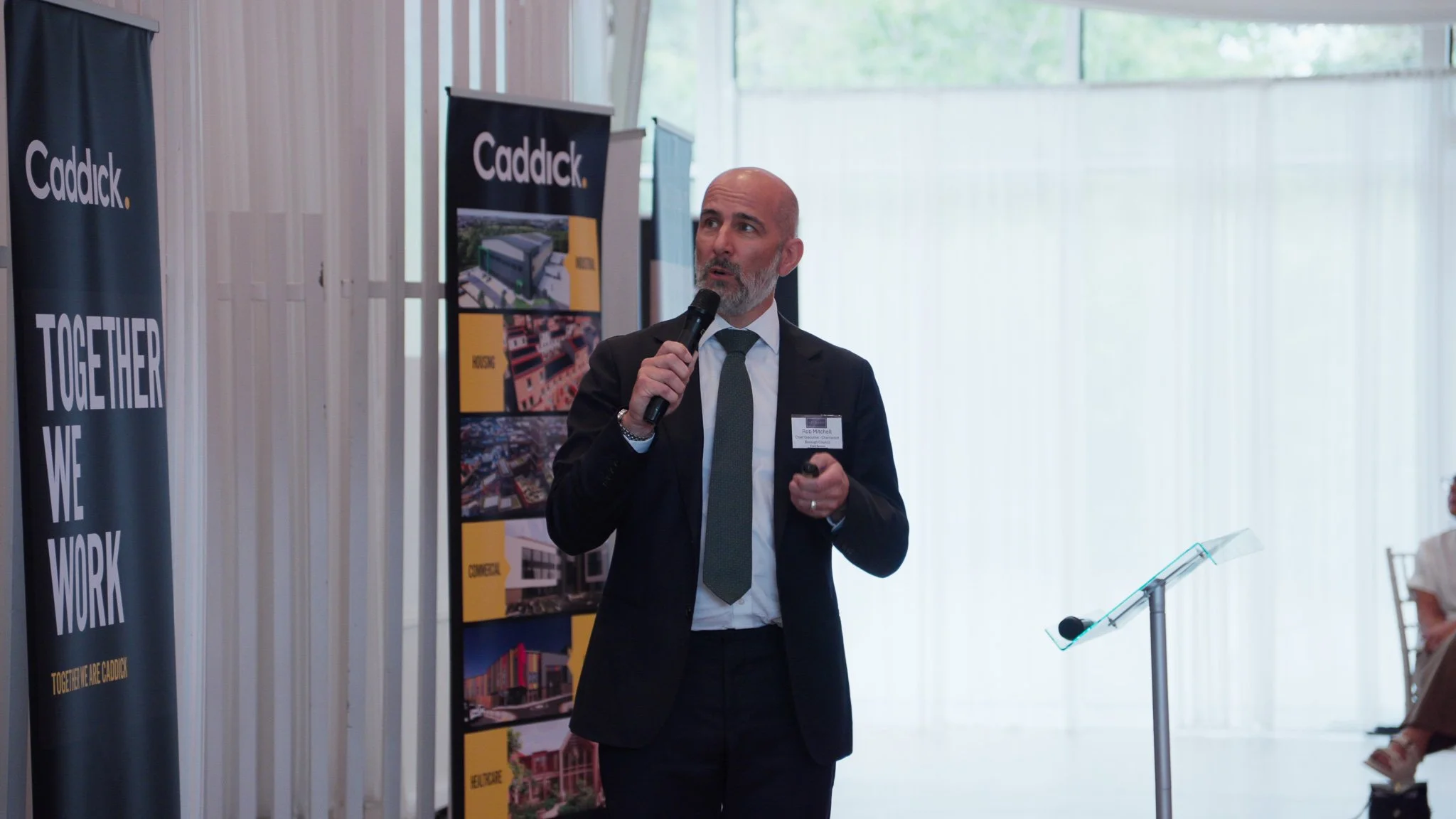 A man in a suit holding a microphone and speaking at an event with banners and a display of architectural designs in the background.