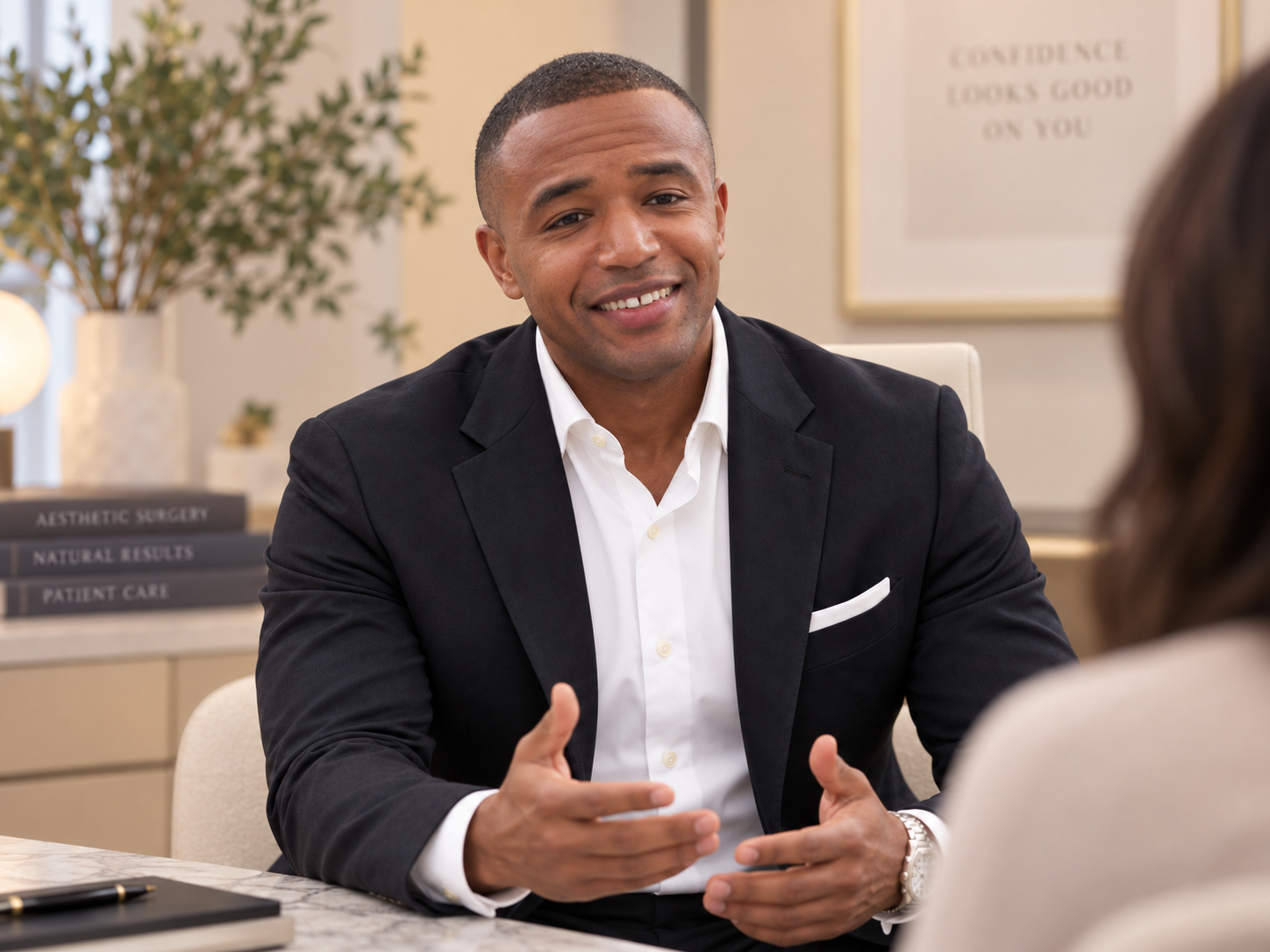 A man in a black suit and white shirt smiling and gesturing during a conversation in an office setting.