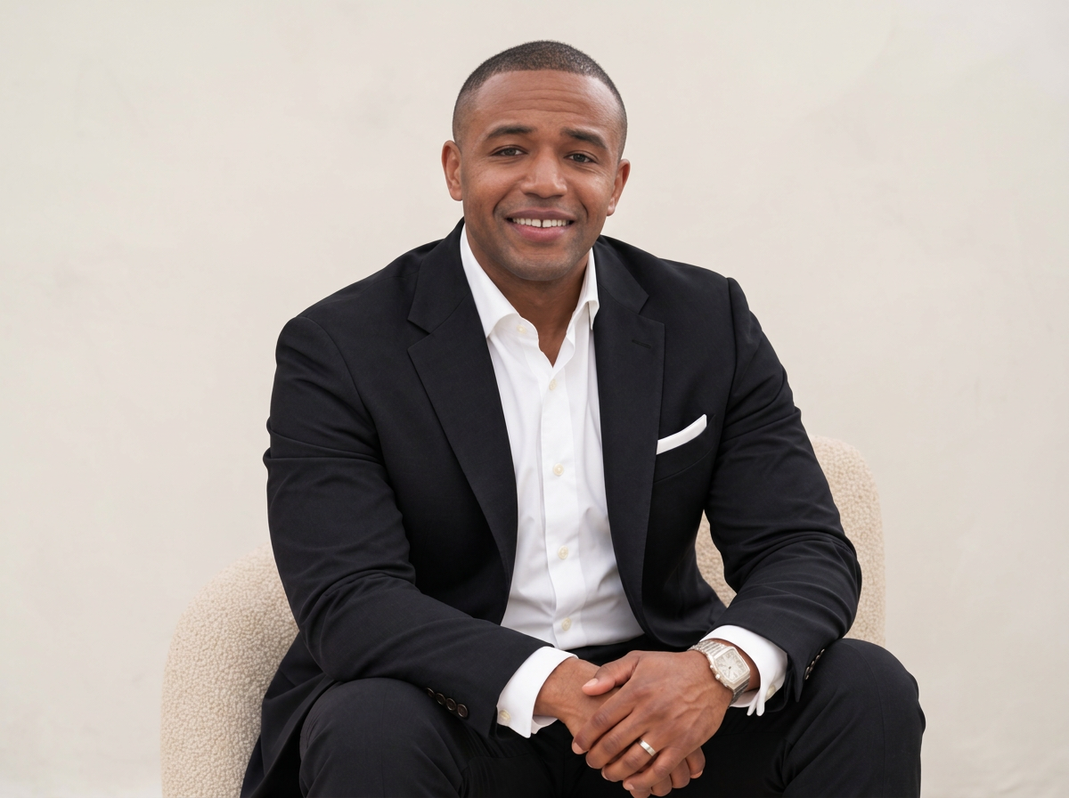 A man dressed in a black suit with a white dress shirt, sitting on a beige chair, smiling with his hands clasped, against a plain light-colored background.