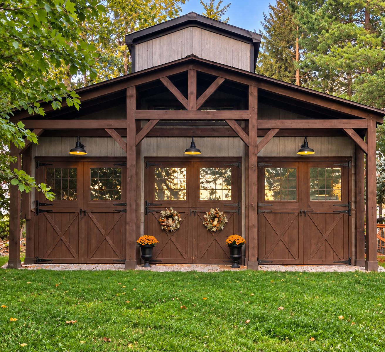 A wooden barn with double doors decorated with fall wreaths, potted orange flowers, and covered in warm yellow string lights, surrounded by green grass and leafy trees.
