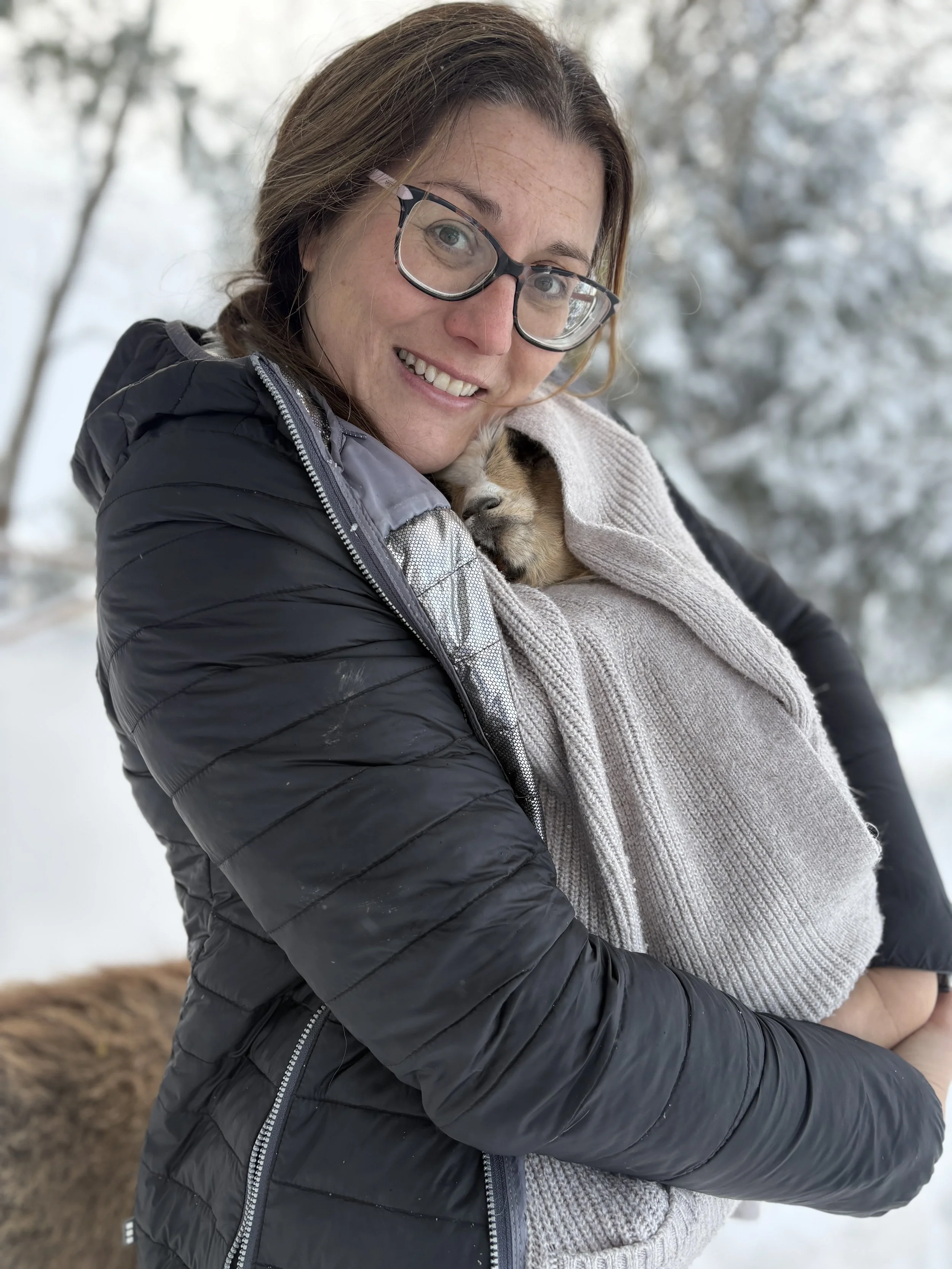 A woman with brown hair, glasses, and a black jacket holding a small puppy wrapped in a beige blanket outdoors in a snowy setting.
