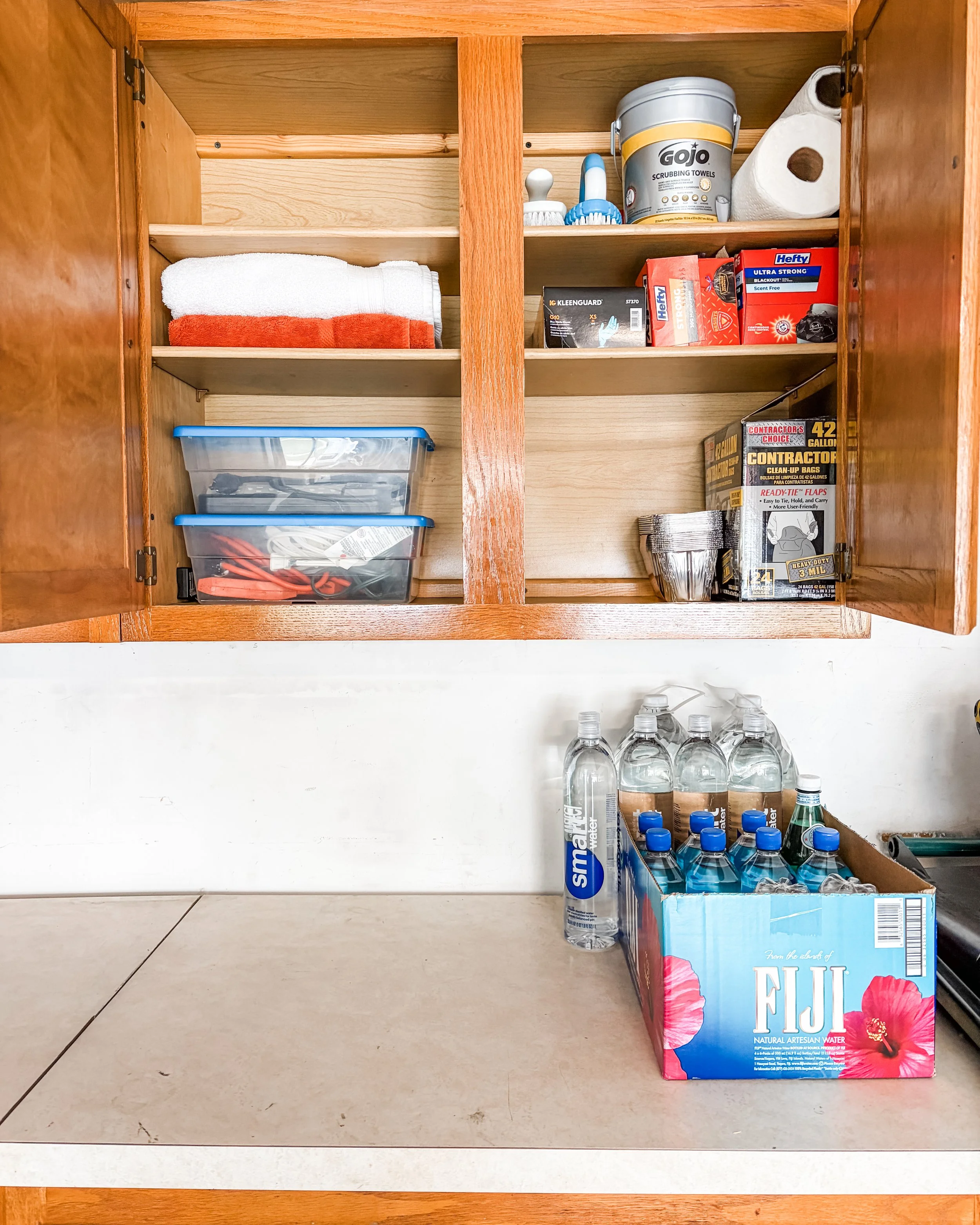 Open kitchen cabinet with various household items, including towels, cleaning supplies, a box of bottled water, and a box of facial tissues. Items are neat, tidy and minimal.