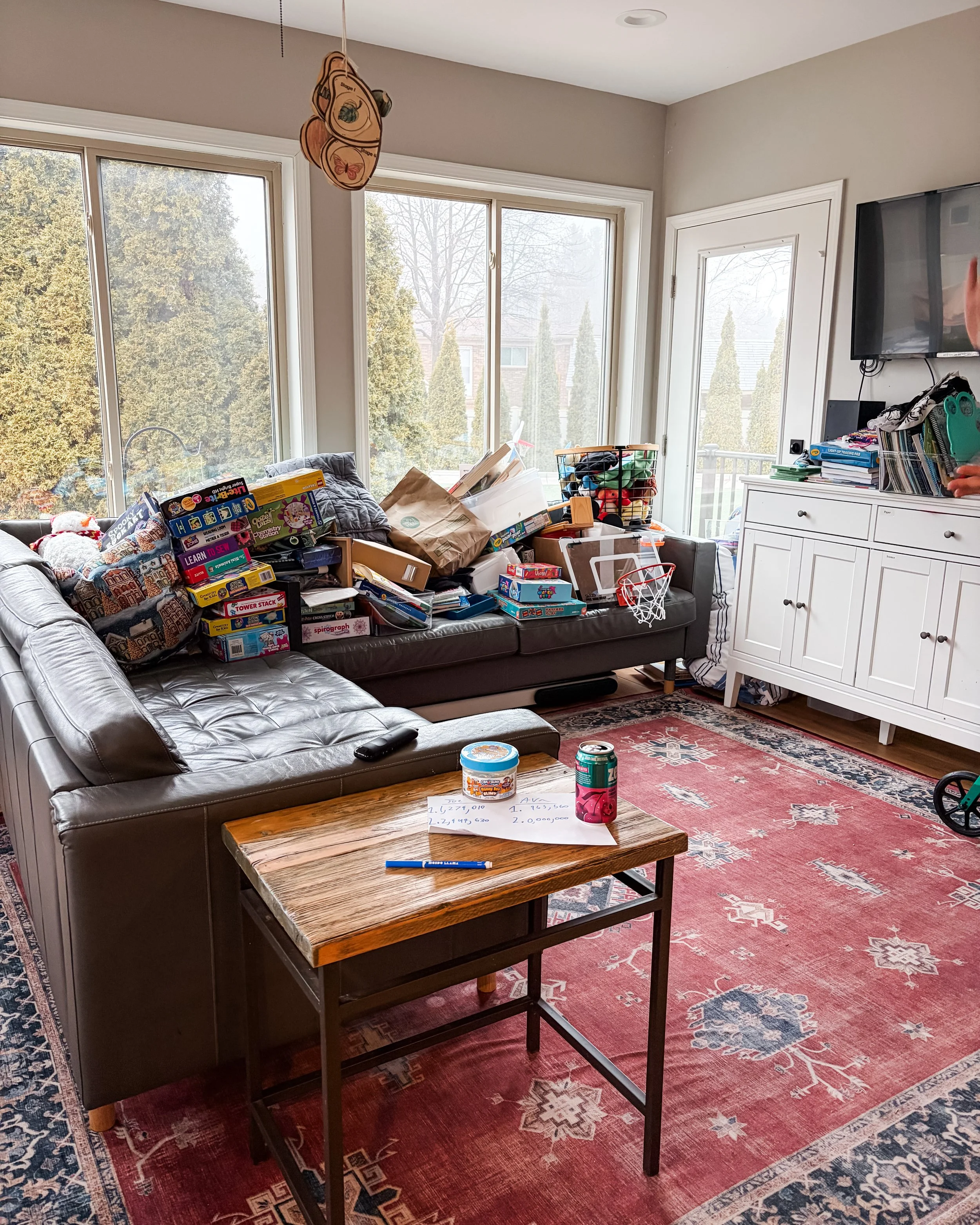 Living room with large windows, cluttered with various items including toys, books, and papers. A leather sofa and a small wooden table with notes and drinks on it, a white cabinet, and a mounted TV.