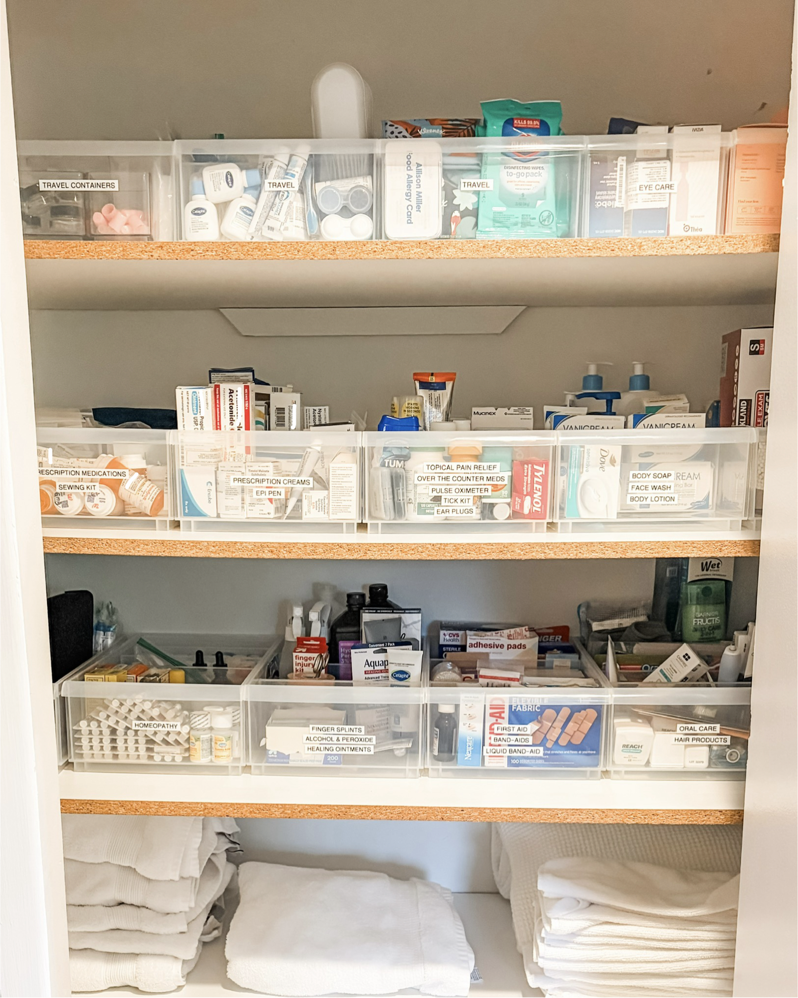 Medical supplies and medications organized in a cabinet with labeled sections such as travel containers, prescription medications, first aid, and oral care, along with neatly folded towels or cloths on the bottom shelf.