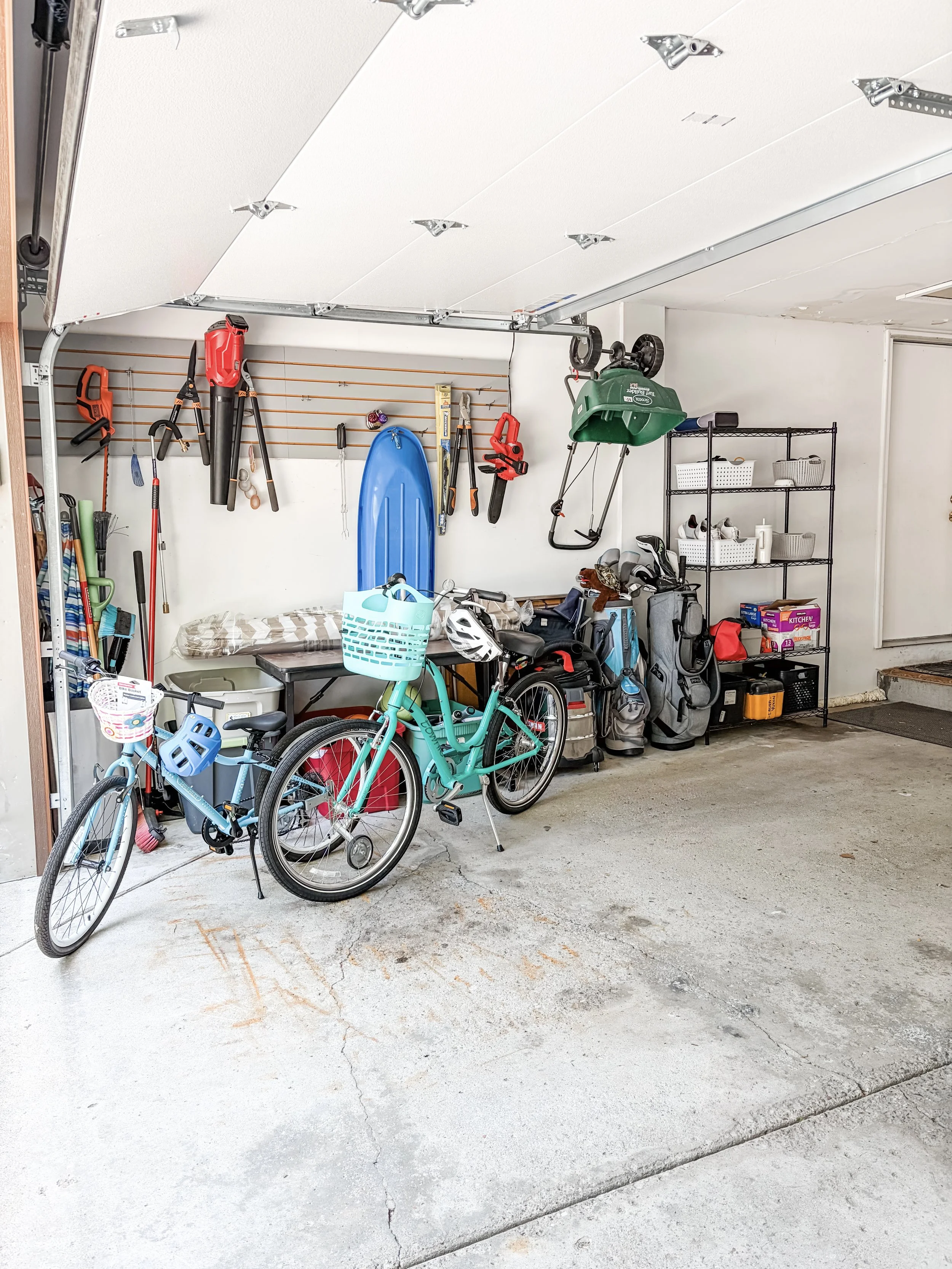 A garage with a bike, gardening tools, and storage shelves. A blue sled hangs on the wall, and a green leaf blower is mounted above the shelves. The concrete floor has some cracks and stains.