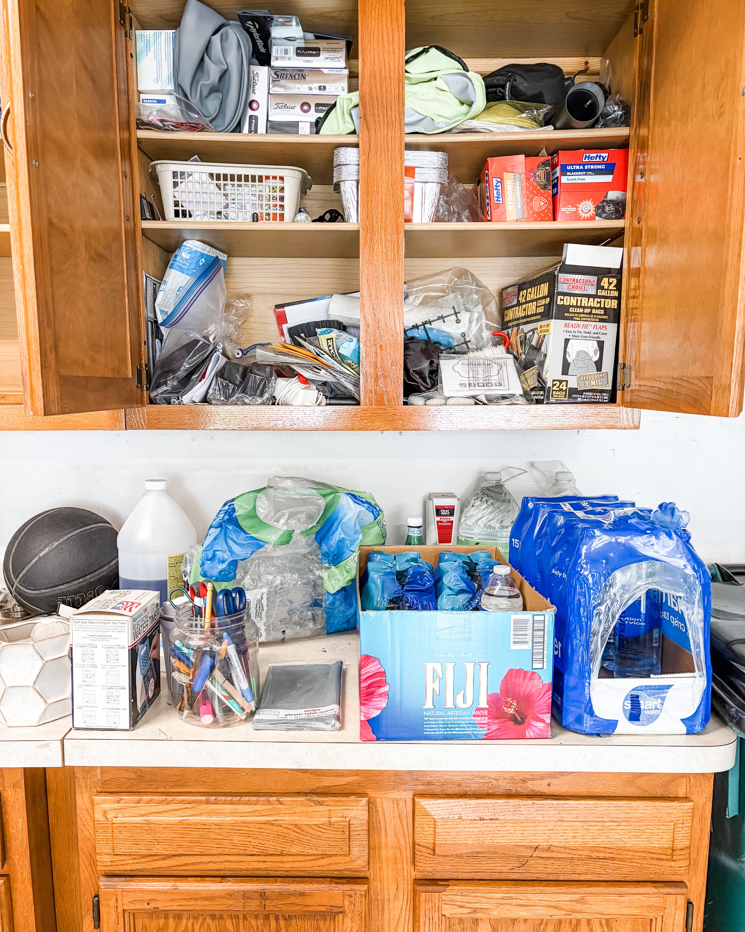 Open kitchen cabinet filled with various items including water bottles, plastic bags, food and drink packages, a box of tissues, small containers, a tablet, and miscellaneous kitchen supplies.